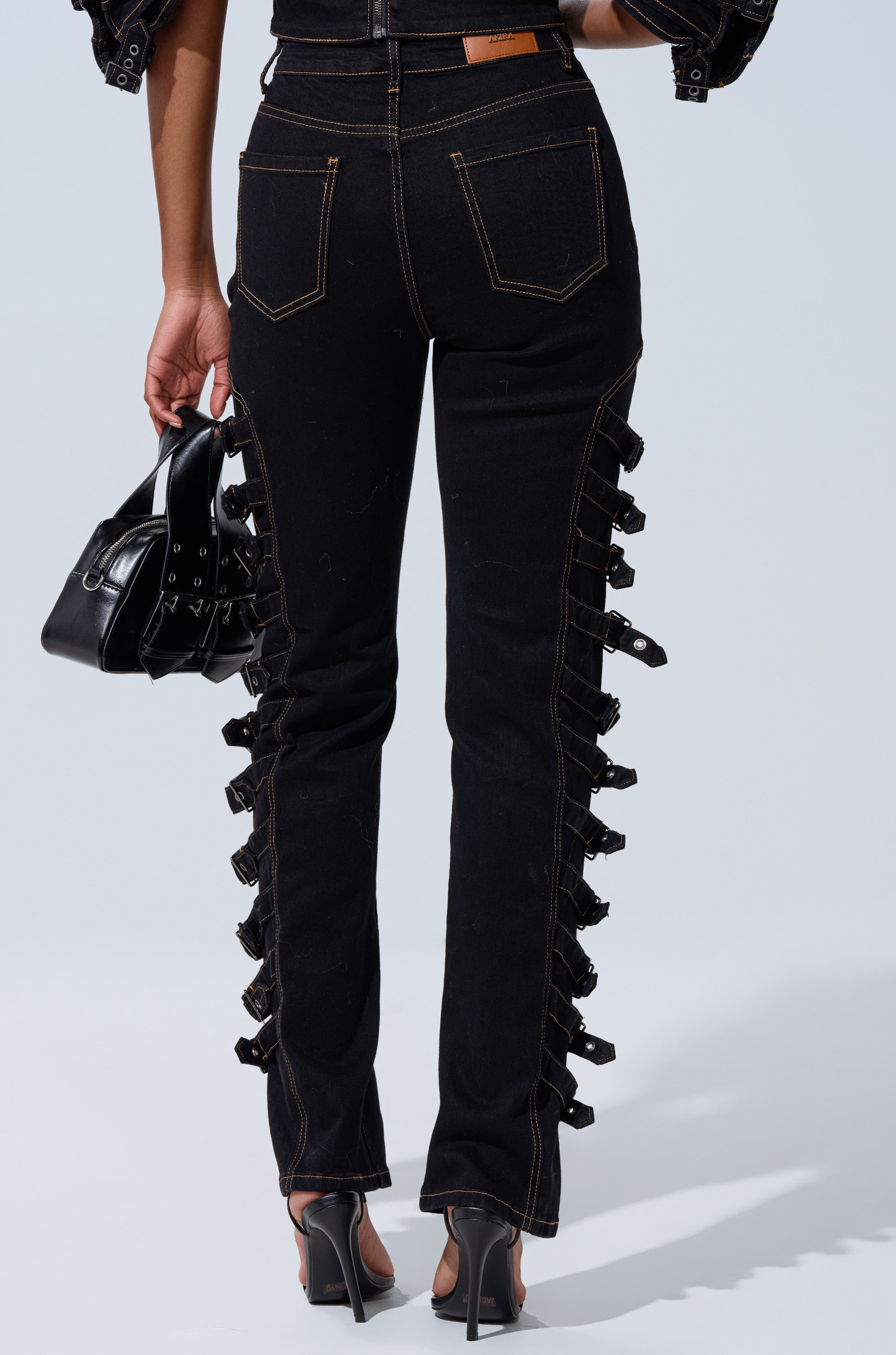 A person wearing the BUCKLE UP SKINNY DENIM PANT, styled with a black handbag and black heeled sandals, photographed from the back.