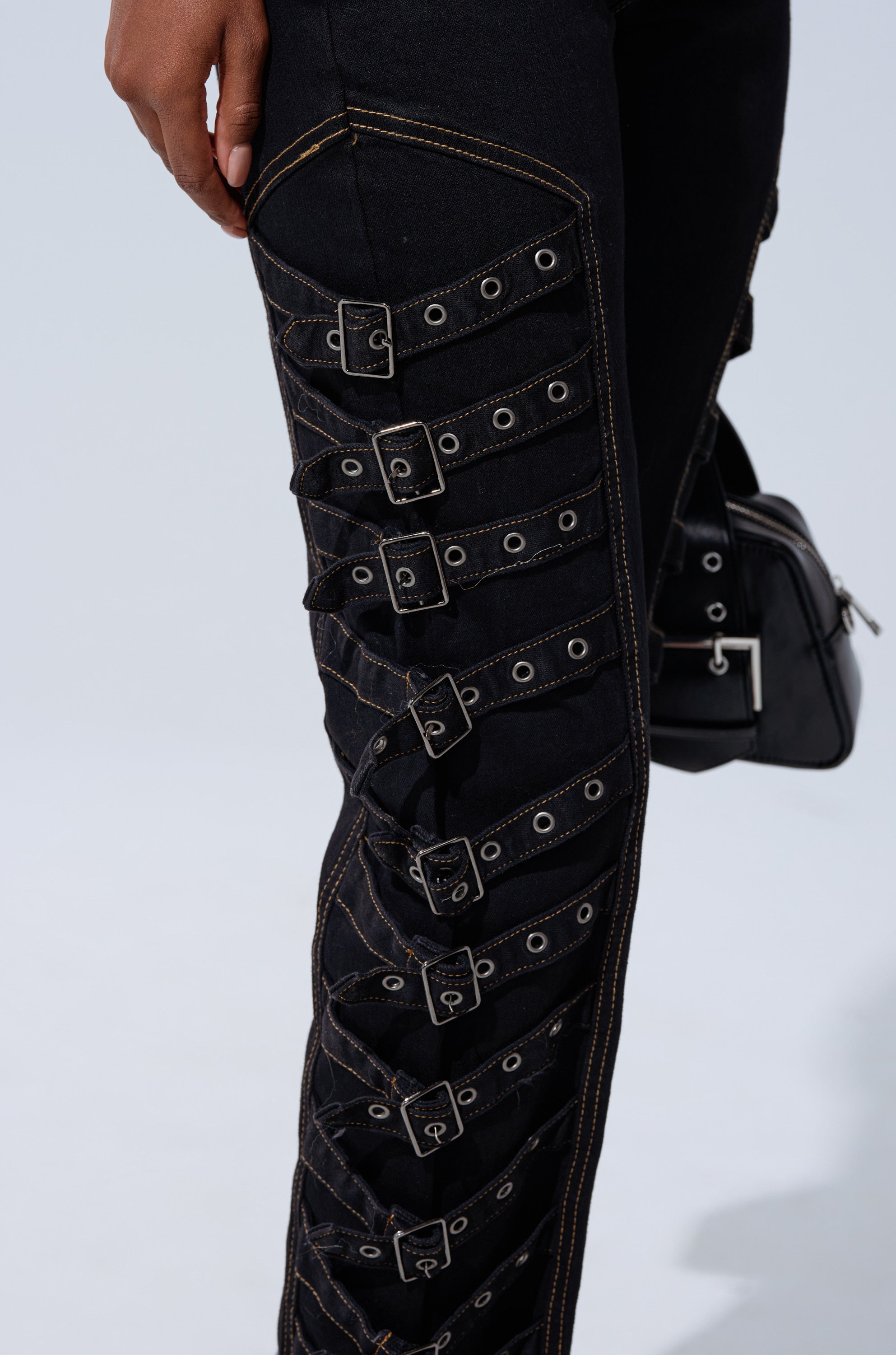 A close-up of a person wearing the BUCKLE UP SKINNY DENIM PANT—high-rise black jeans with multiple horizontal buckles, grommets along the thigh and knee, contrast stitching, and a small black handbag in the background.