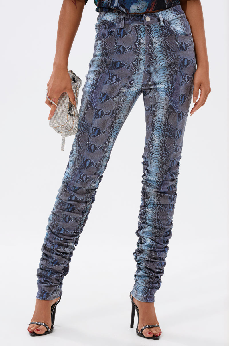 A person wears the DAYS END RUCHED STACKED SNAKE SKINNY DENIM in blue and black, paired with black high-heeled sandals and a sparkly silver clutch. The photo is cropped from the shoulders down. 0