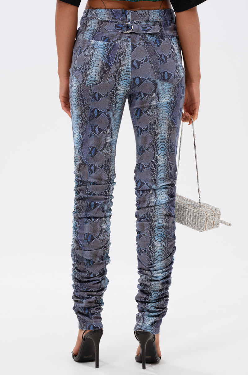 A person stands facing away on a plain white background, wearing the DAYS END RUCHED STACKED SNAKE SKINNY DENIM in blue and gray snakeskin print with black high heels and holding a small silver handbag. 2