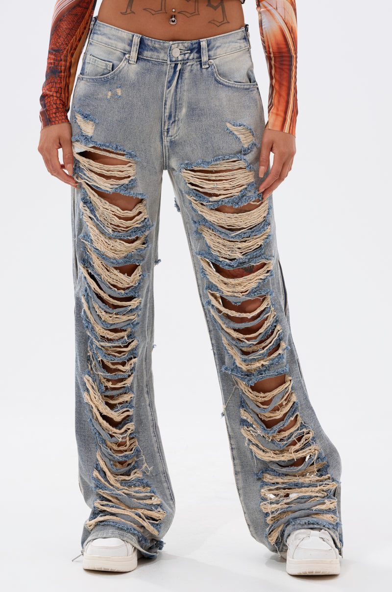 The model wears REAL TALK DISTRESSED JEANS in vintage-wash blue with large rips, styled with white sneakers and a patterned long-sleeve crop top, standing against a plain background. 0