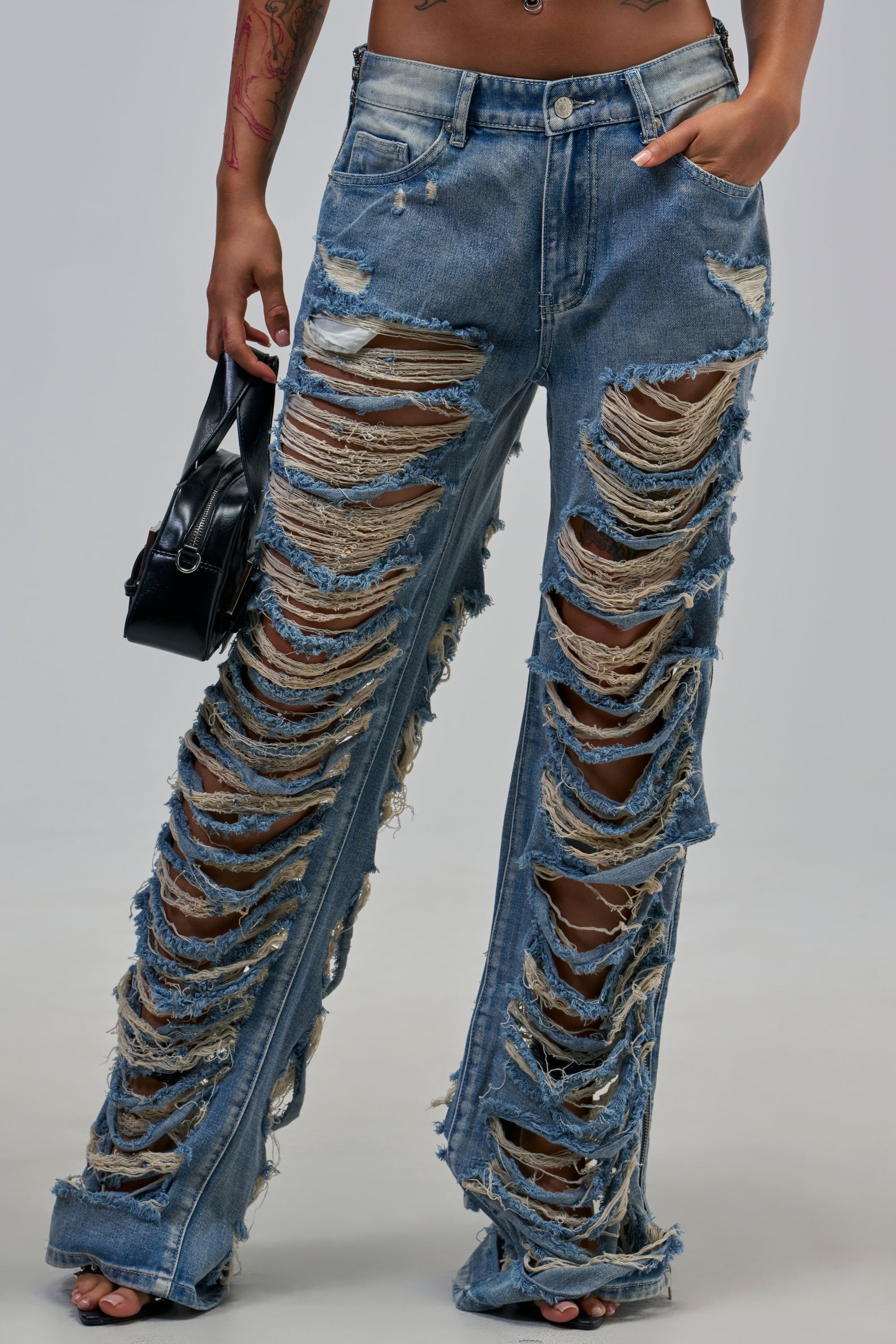 Model wears REAL TALK DISTRESSED JEANS with large rips, open-toe shoes, and a black handbag for a bold street style. The light blue denim features heavy distressing on both legs against a neutral backdrop.