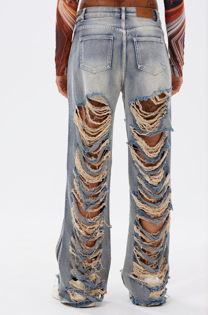 A person models the REAL TALK DISTRESSED JEANS in light blue vintage wash, featuring large horizontal rips and frayed edges on the back of both legs, standing against a plain white background. 3