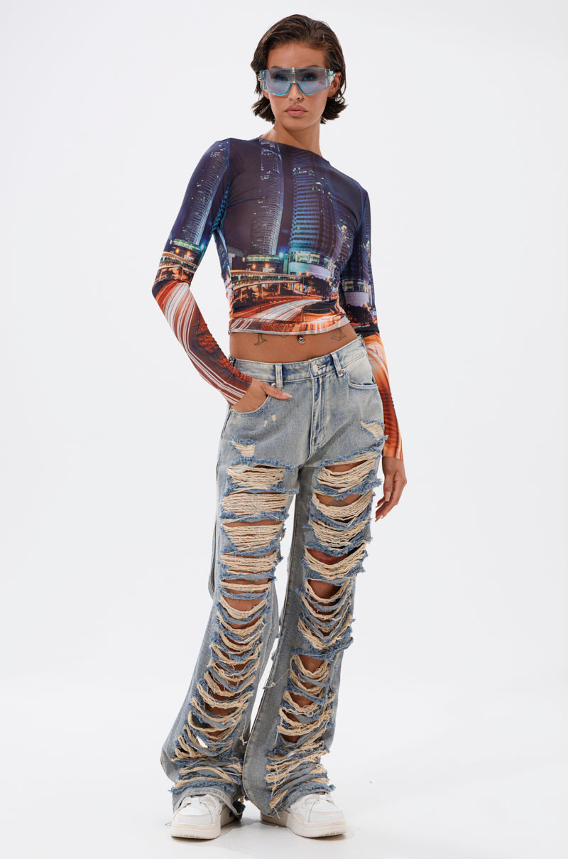 Person standing against a white background, wearing blue sunglasses, a long-sleeve cityscape print crop top, and REAL TALK DISTRESSED JEANS styled with white sneakers. 4