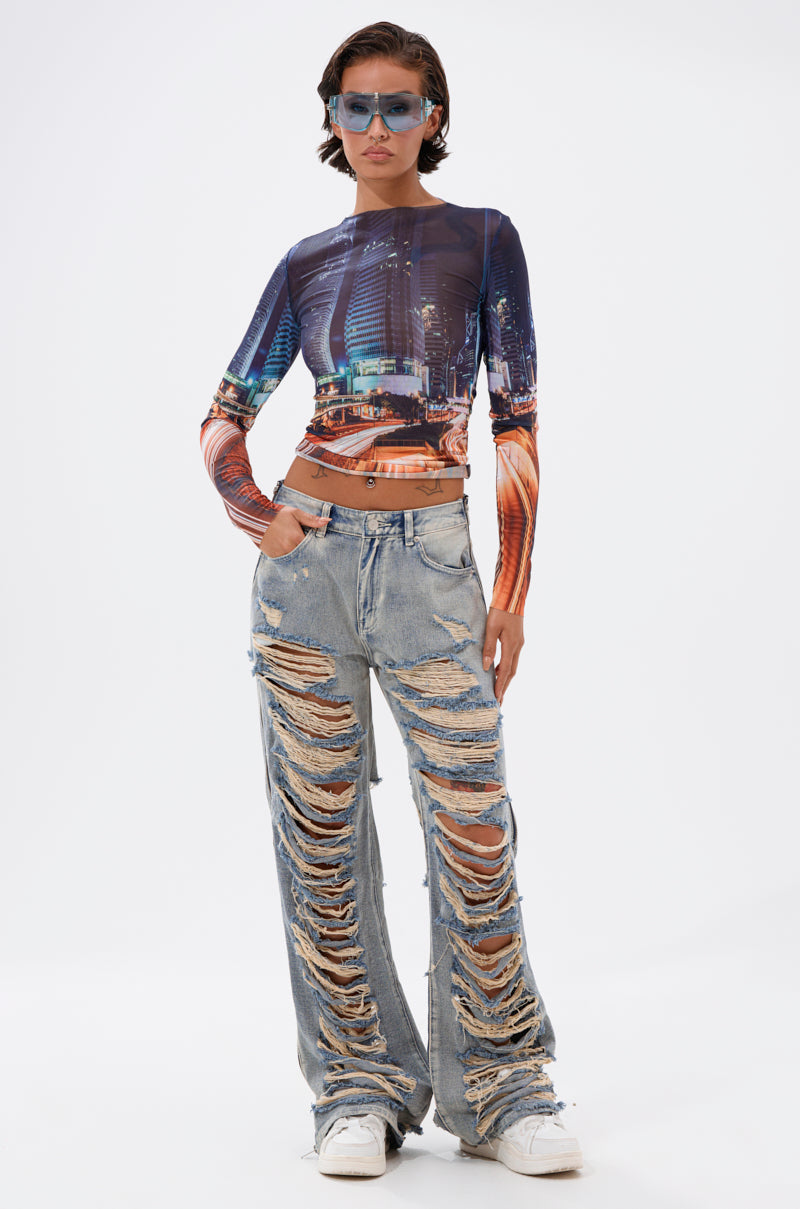 A person stands confidently against a white background wearing blue sunglasses, a cityscape print long-sleeve crop top, and REAL TALK DISTRESSED JEANS. 5