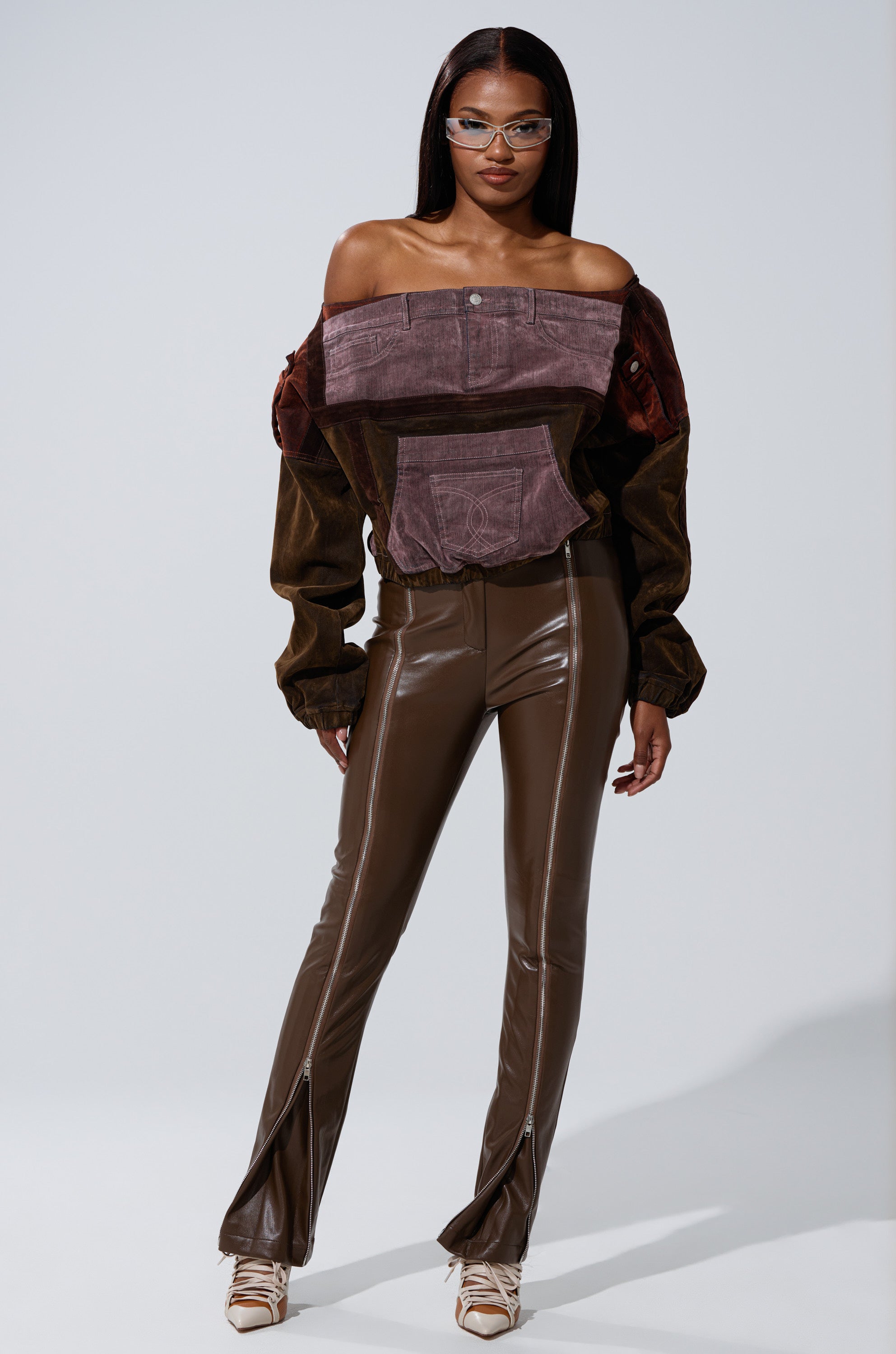A woman in glasses, an off-shoulder brown and purple top, FEELING THE VIBES FAUX LEATHER LEGGING with side hem slits, and beige high heels poses against a plain gray background. 7