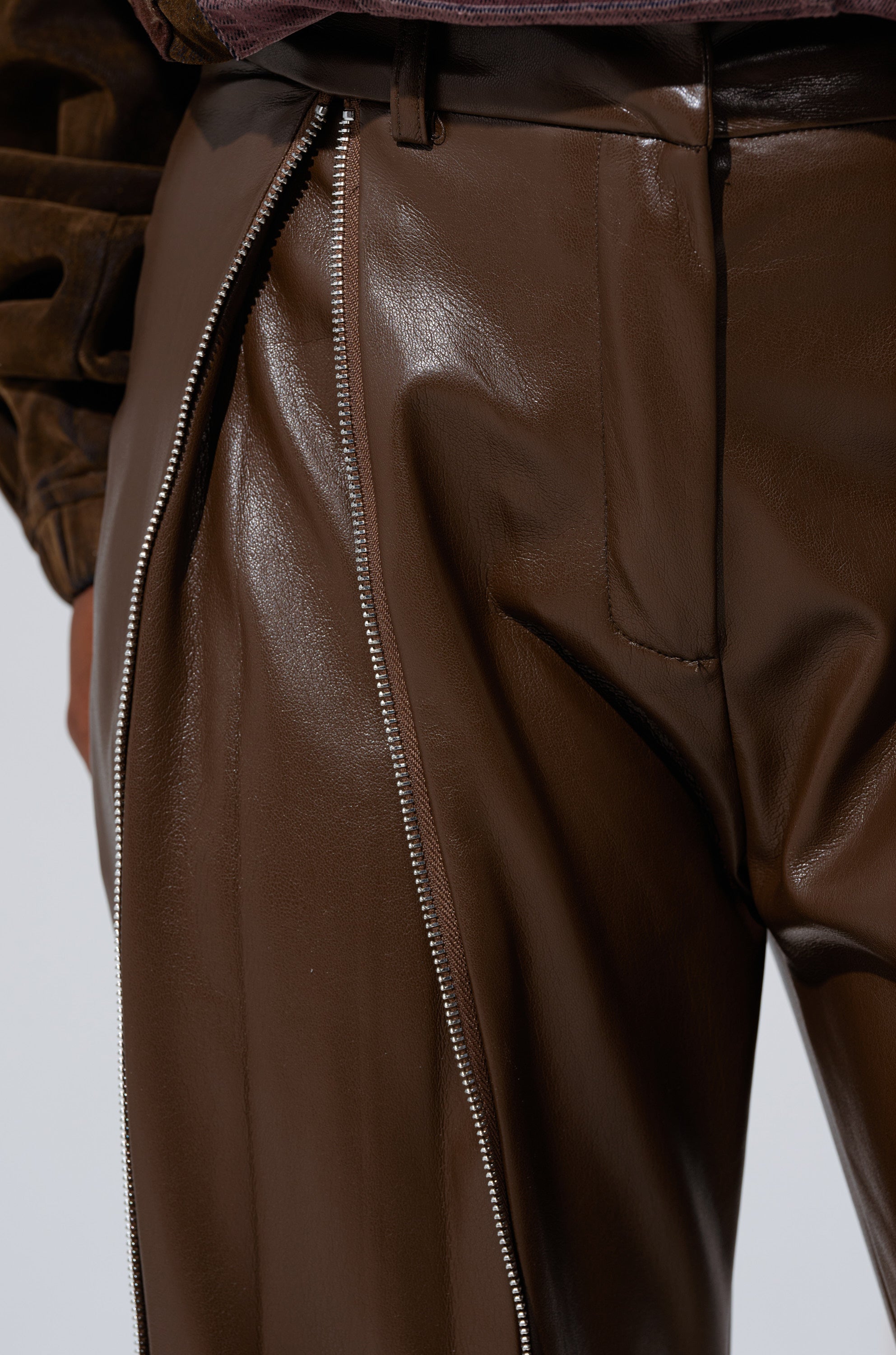 Close-up of the FEELING THE VIBES FAUX LEATHER LEGGING in glossy brown, featuring a large diagonal silver zipper partially open to reveal matching brown fabric, with focus on the waistband and upper leg area. 9