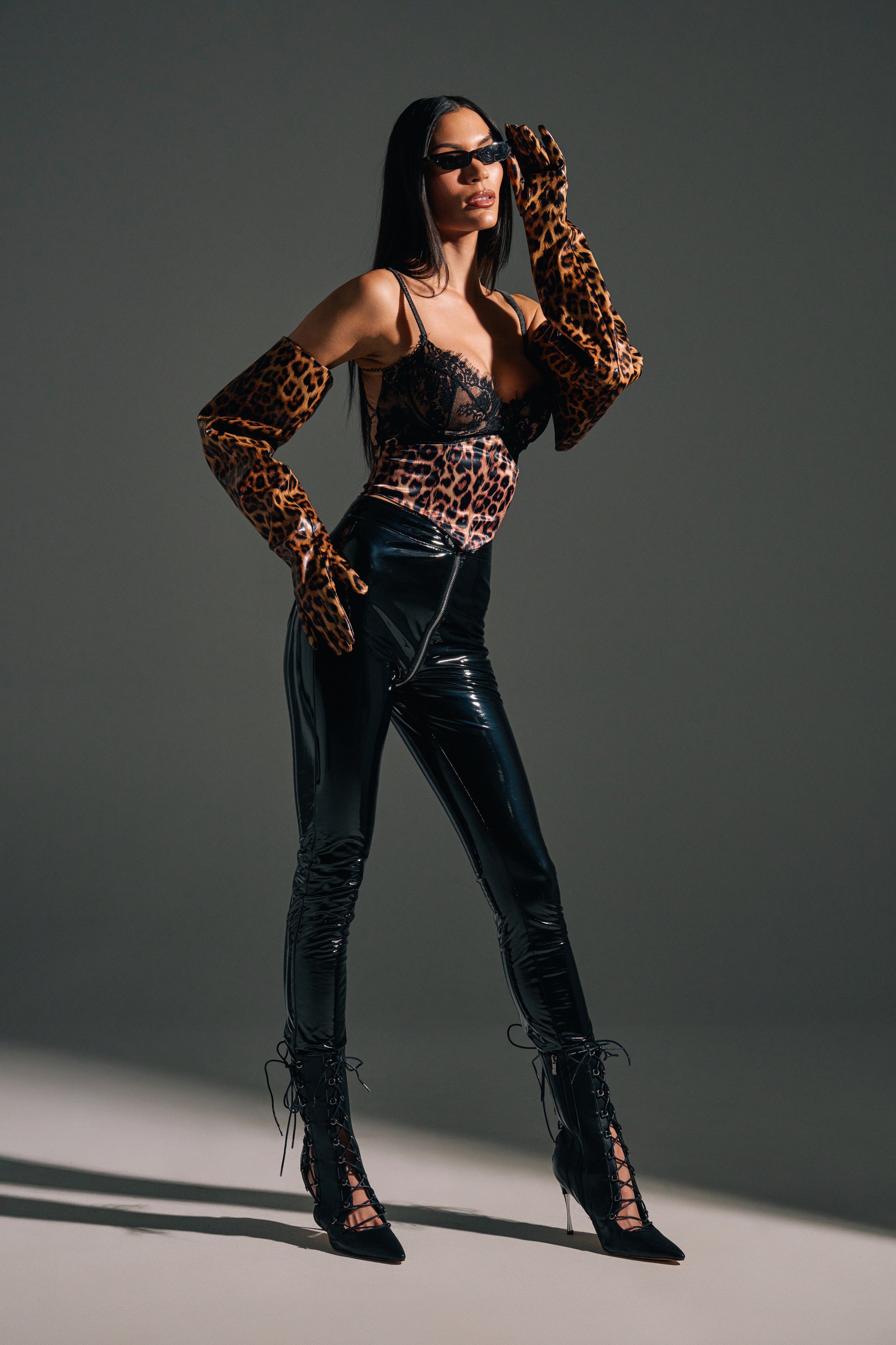 A woman poses confidently in the DROP IT LIKE ITS HOT VINYL LEGGING WITH ZIPPER, paired with a lacy black corset top, leopard-print gloves, lace-up heels, and sunglasses against a plain gray background.