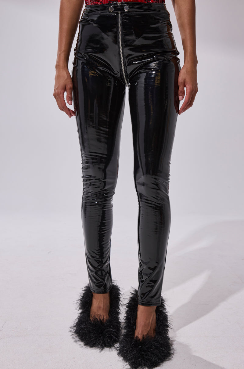 A person is shown from the shoulders down, wearing DROP IT LIKE ITS HOT VINYL LEGGING WITH ZIPPER and black fuzzy slippers, hands by their sides against a plain background.