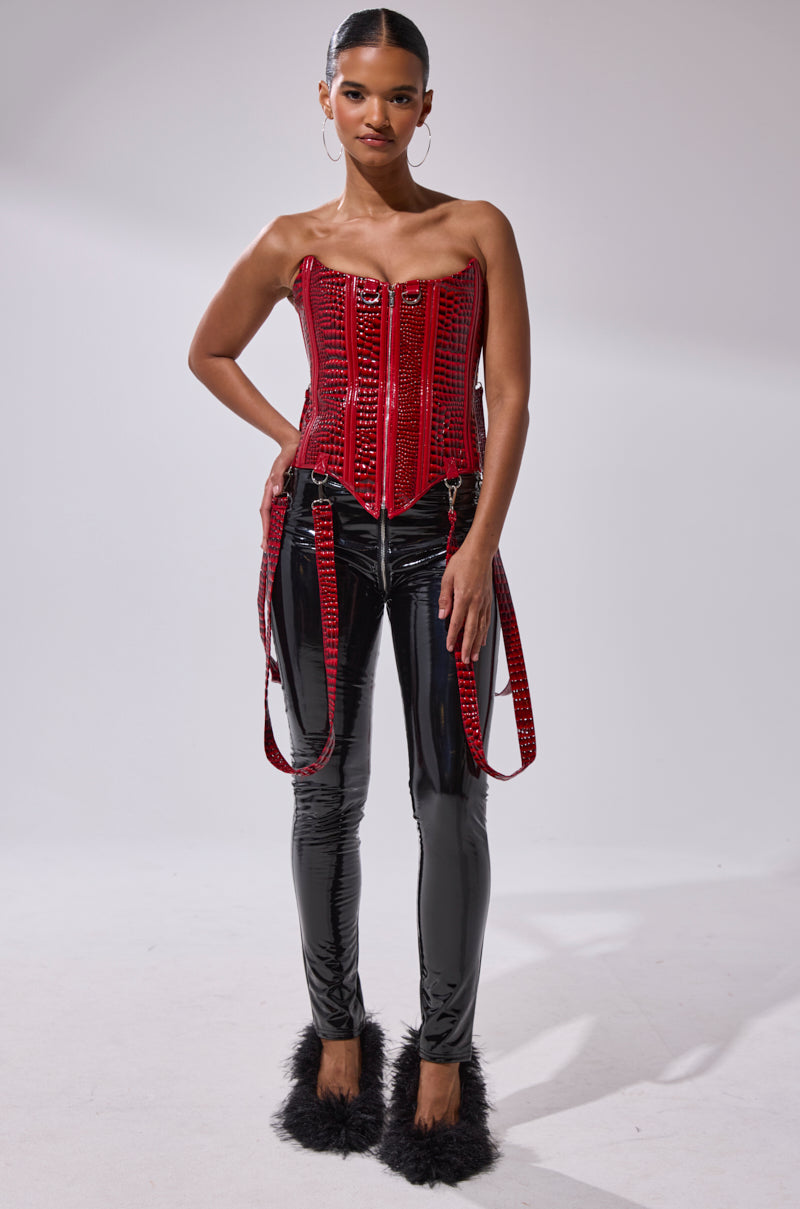A woman stands confidently in a shiny red corset, DROP IT LIKE ITS HOT VINYL LEGGING WITH ZIPPER, and black fuzzy high heels against a plain light background.