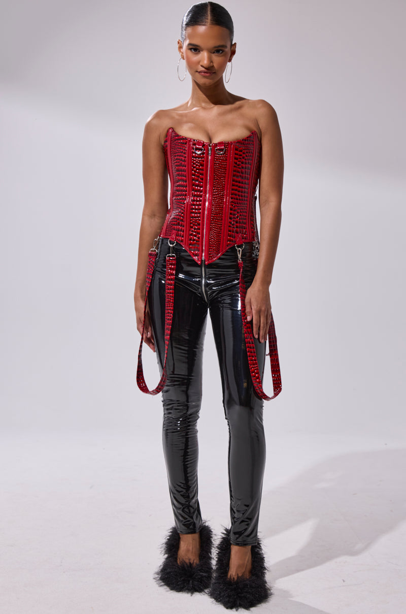 A woman stands against a plain background wearing a shiny red strapless corset, hoop earrings, black furry heels, and the DROP IT LIKE ITS HOT VINYL LEGGING WITH ZIPPER.
