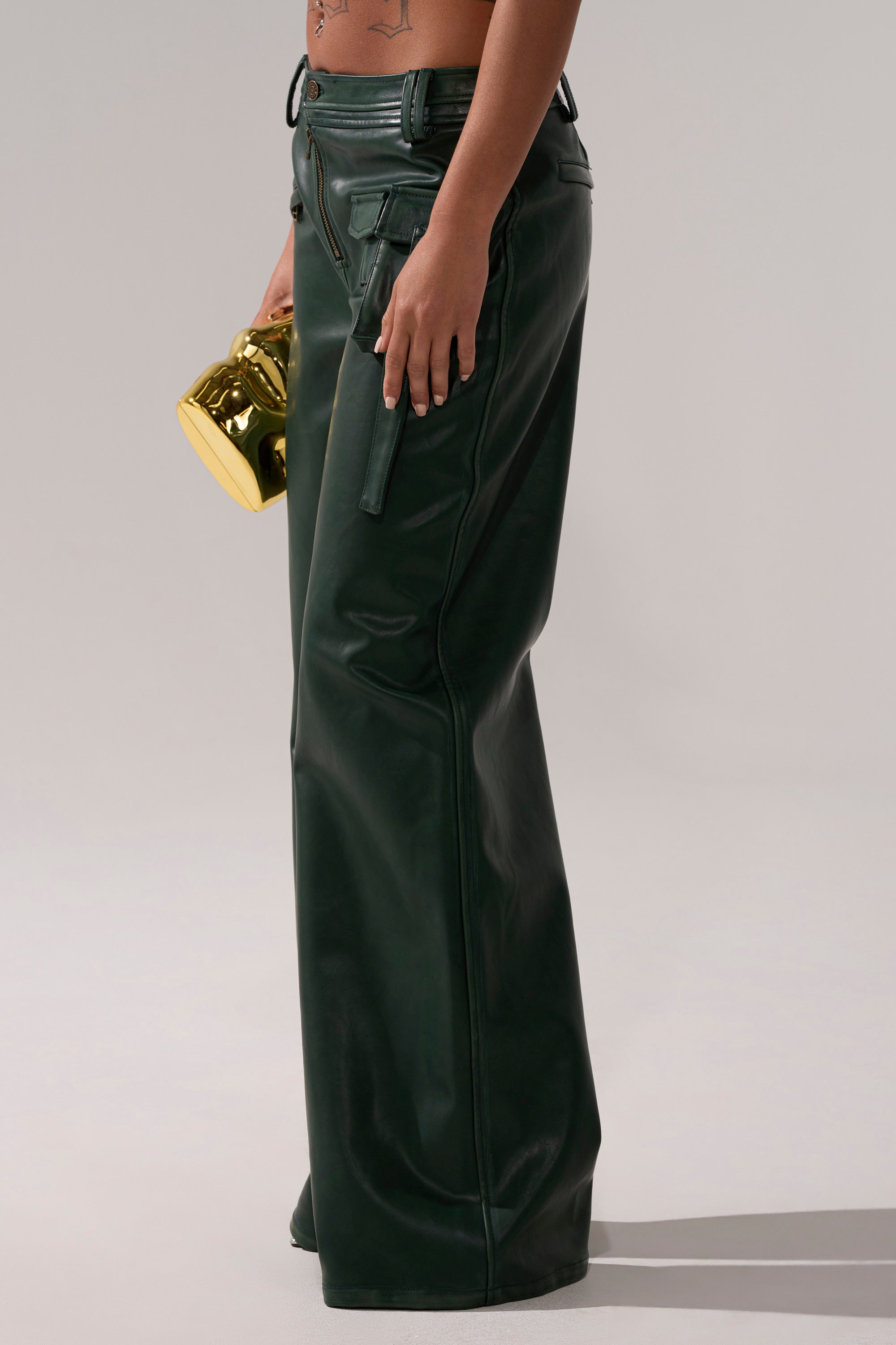 A person wearing SO YOU THOUGHT TROUSER in dark green faux leather stands sideways, holding a small gold object. Their bare torso reveals tattoos on their side and lower stomach against a plain light grey background. 3