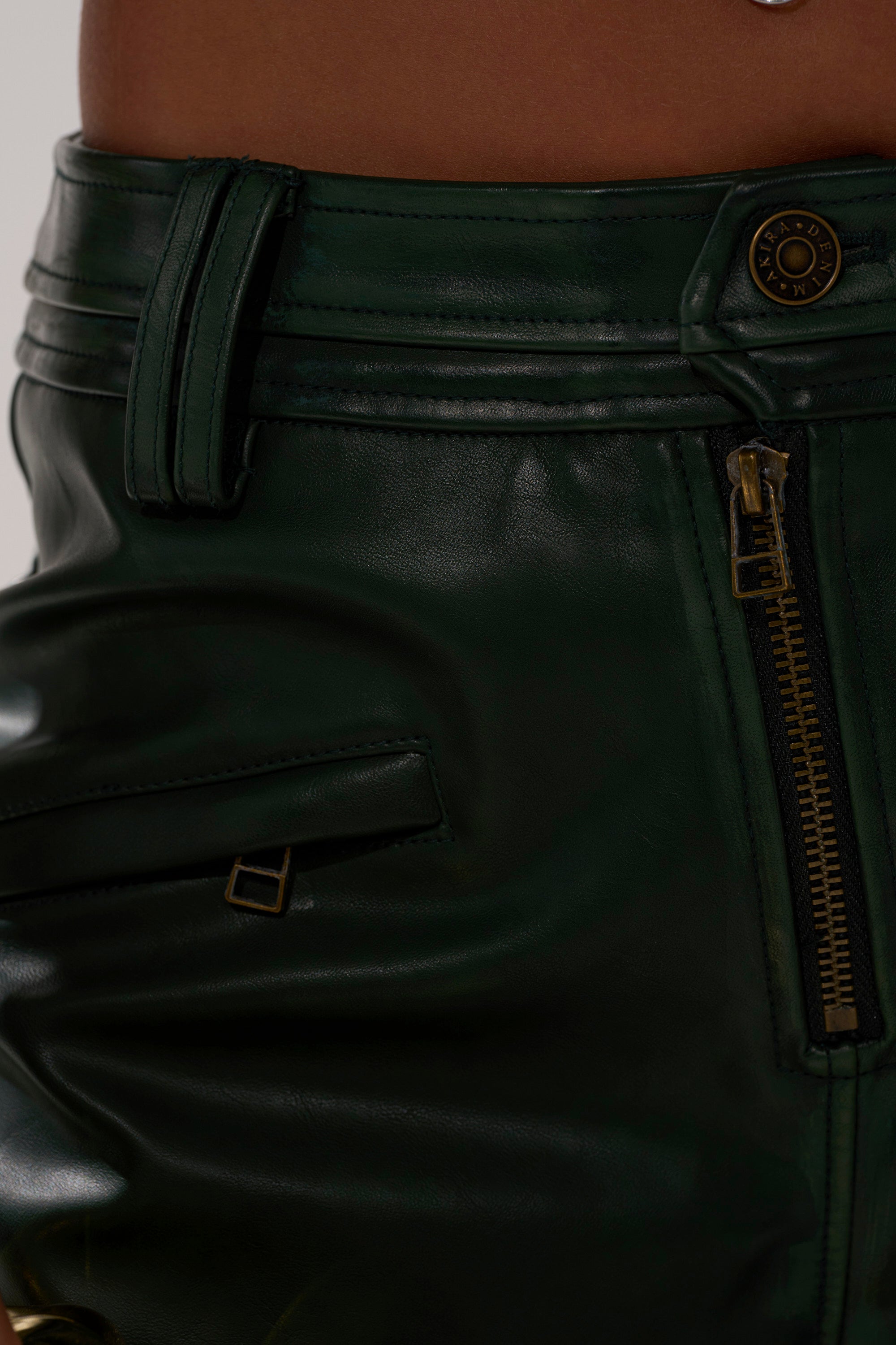 Close-up of the SO YOU THOUGHT TROUSER in dark green faux leather, featuring a zipper, button closure, belt loops, and a horizontal pocket with a small metal detail. The image captures part of a person's lower torso. 7