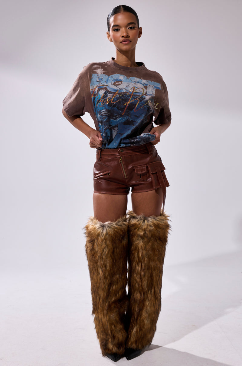 A woman with sleek hair and a confident look wears a graphic tee, the NICOLETTE FAUX LEATHER SHORT featuring cargo pockets, and tall furry brown leg warmers against a plain background. 2