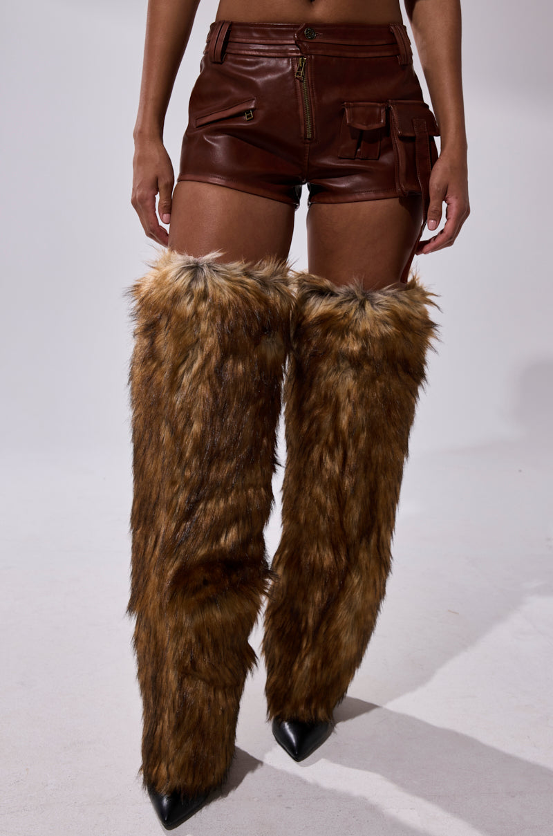 A person wears the NICOLETTE FAUX LEATHER SHORT with zipper and pocket details, styled with tall furry brown leg warmers and black pointed-toe shoes, standing against a plain light background. 3