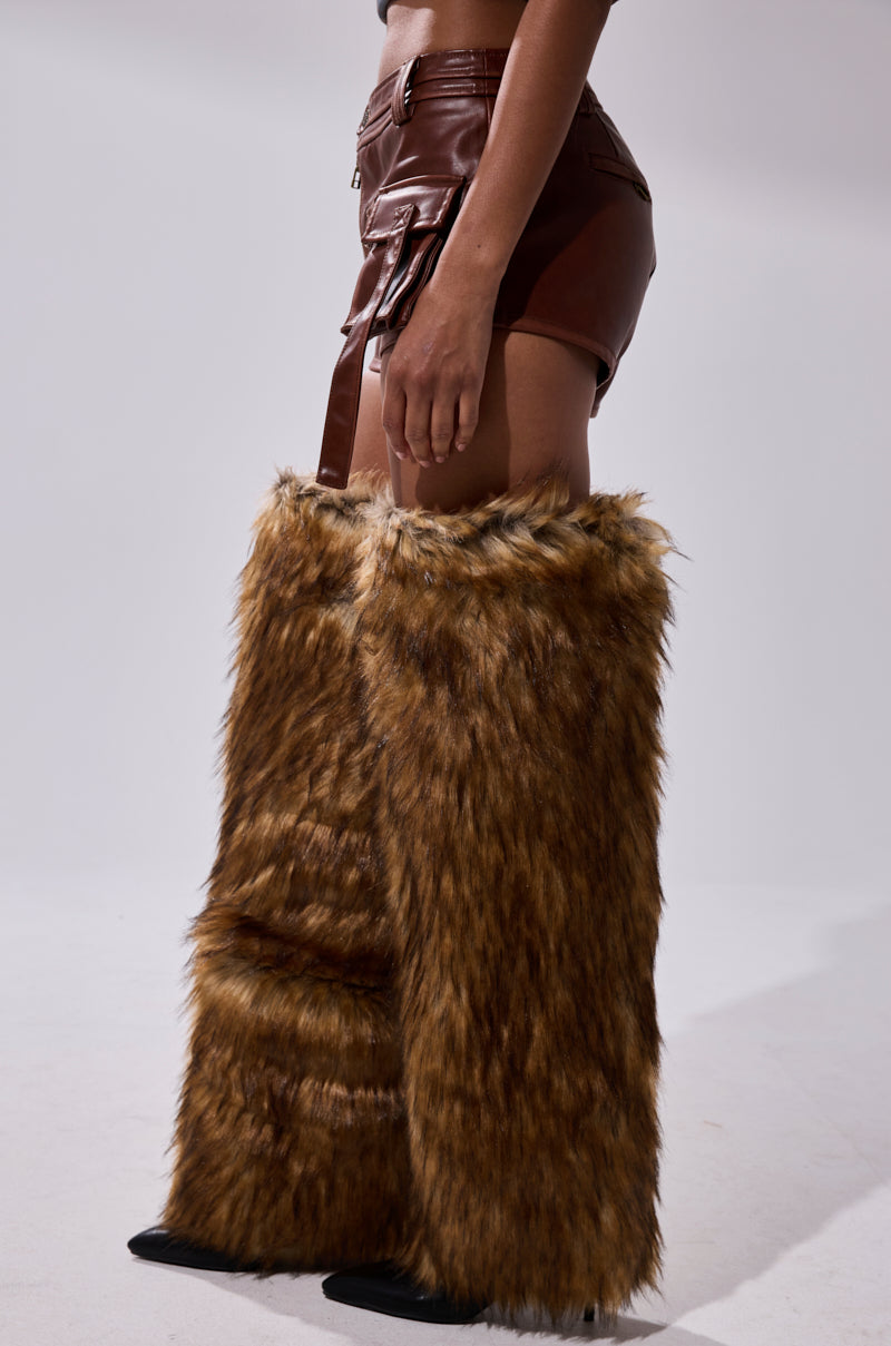 A person stands sideways against a plain light background wearing the NICOLETTE FAUX LEATHER SHORT in brown mini-length and tall, furry brown leg warmers that cover most of their legs. 4