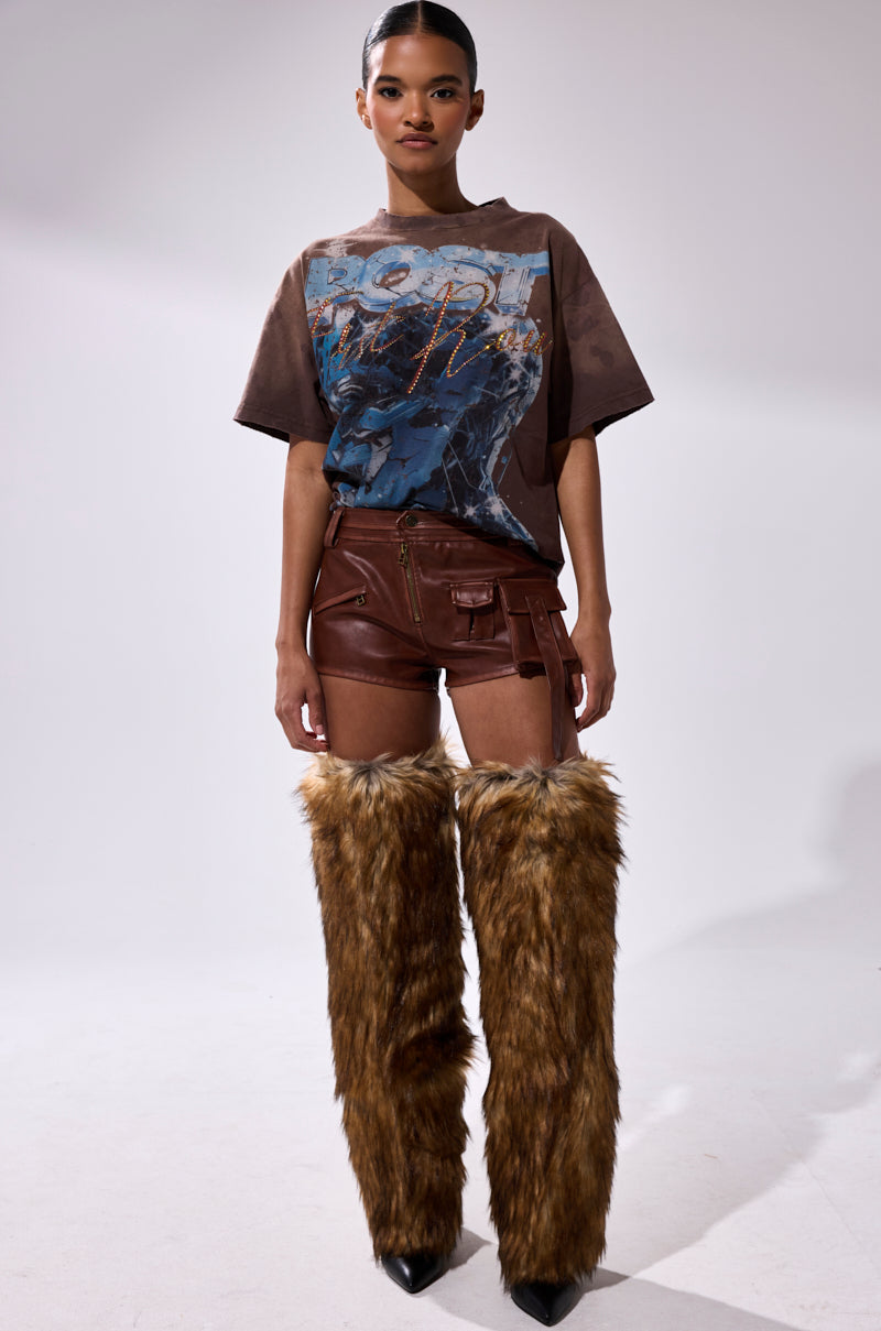 A woman stands against a plain background wearing a loose brown graphic t-shirt, NICOLETTE FAUX LEATHER SHORTS with cargo pockets, and tall brown faux fur leg warmers covering her legs from mid-thigh to feet. 6