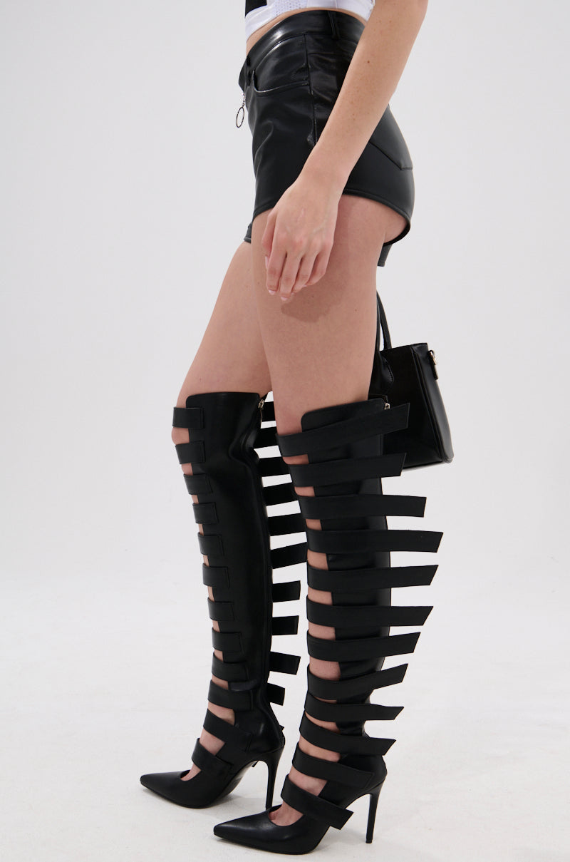 Wearing LOOKS GREAT FAUX LEATHER SHORT and distinctive black thigh-high boots with multiple horizontal cutouts, this person carries a small black handbag. 2
