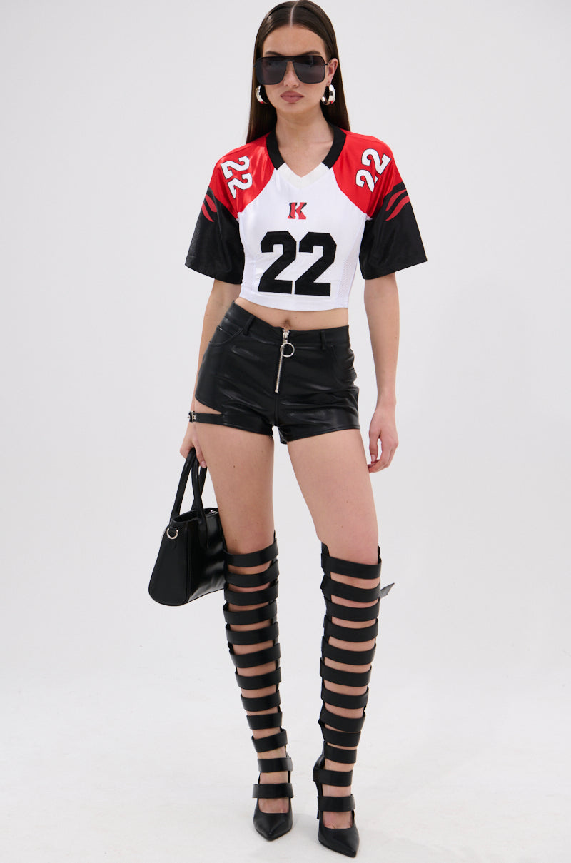 A woman wearing the LOOKS GREAT FAUX LEATHER SHORT, a sporty crop top with 22, thigh-high strappy boots, sunglasses, and hoop earrings stands holding a black handbag against a plain white background. 4