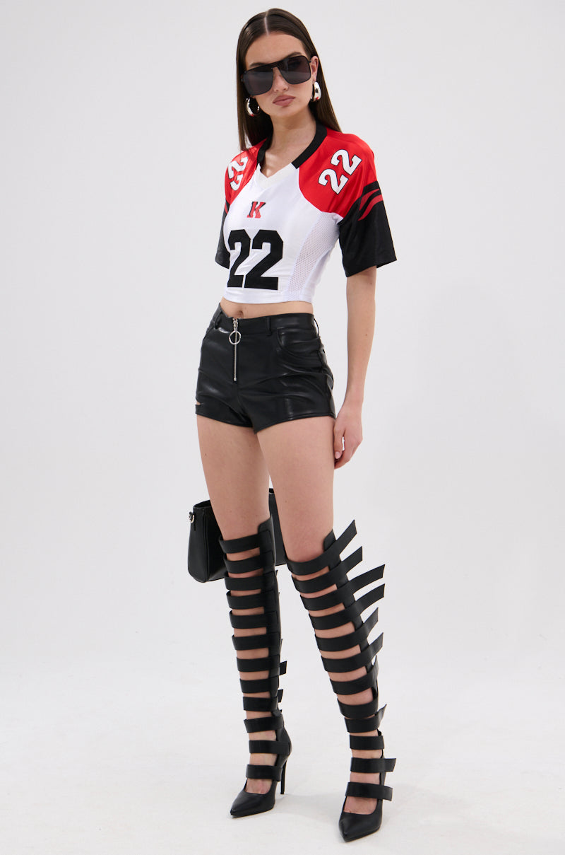 A woman models the LOOKS GREAT FAUX LEATHER SHORT with a red, white, and black sports jersey crop top, black strappy knee-high heeled boots, sunglasses, and hoop earrings against a plain white background. 5