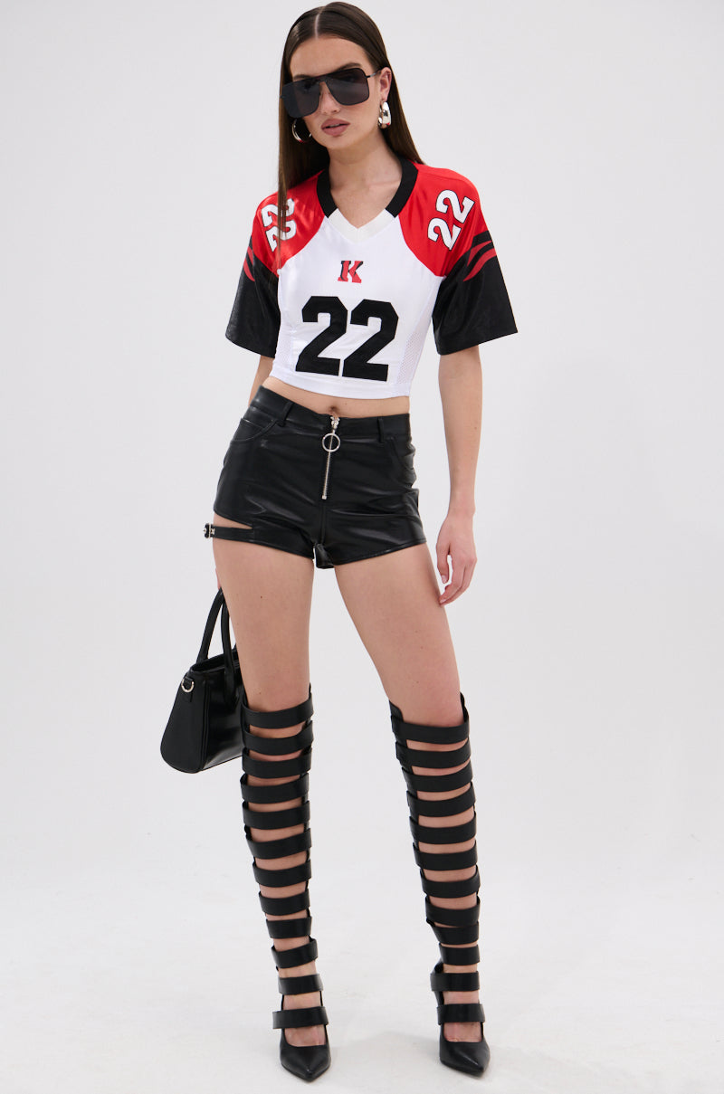 A woman confidently stands against a plain white background wearing LOOKS GREAT FAUX LEATHER SHORTS, a cropped sports jersey, caged thigh-high boots, sunglasses, earrings, and carrying a black handbag. 6