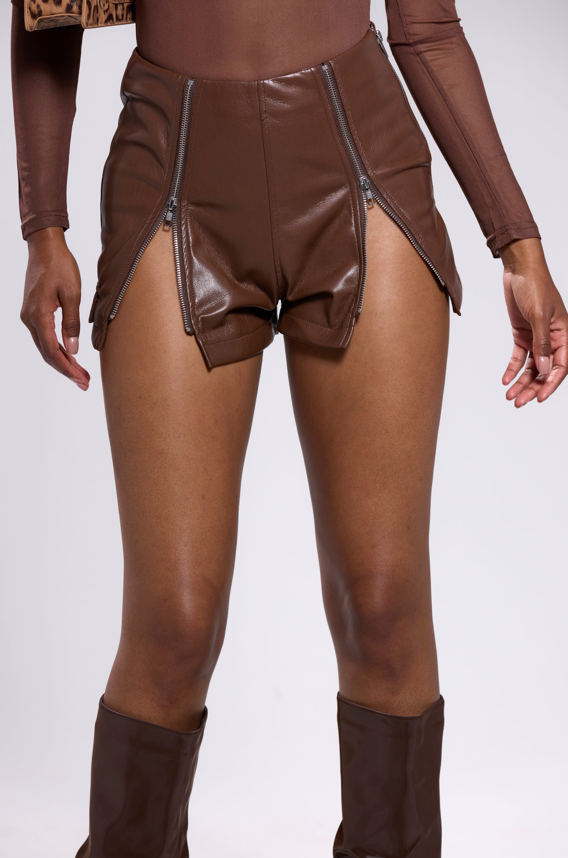 Someone wearing the OH HOT DAMN FAUX LEATHER SHORT WITH ZIPPERS IN BROWN, paired with a matching long-sleeve top and brown knee-high boots, stands with hands by their sides. 8