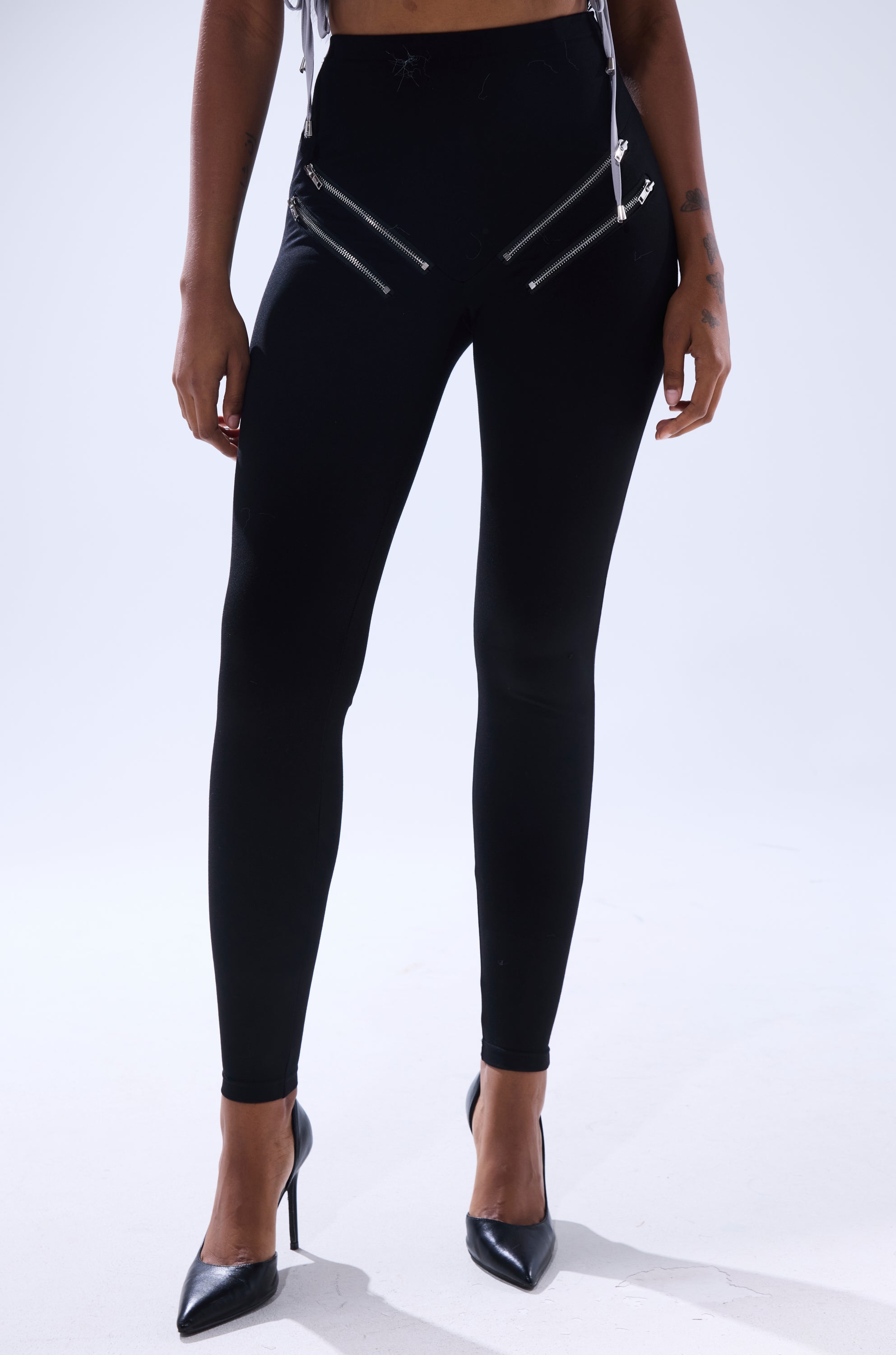 Wearing the ZIP ME UP SEAMLESS LEGGING—black high-rise style with diagonal silver zippers near the hips—paired with black pointed-toe heels, this person stands against a plain light background.