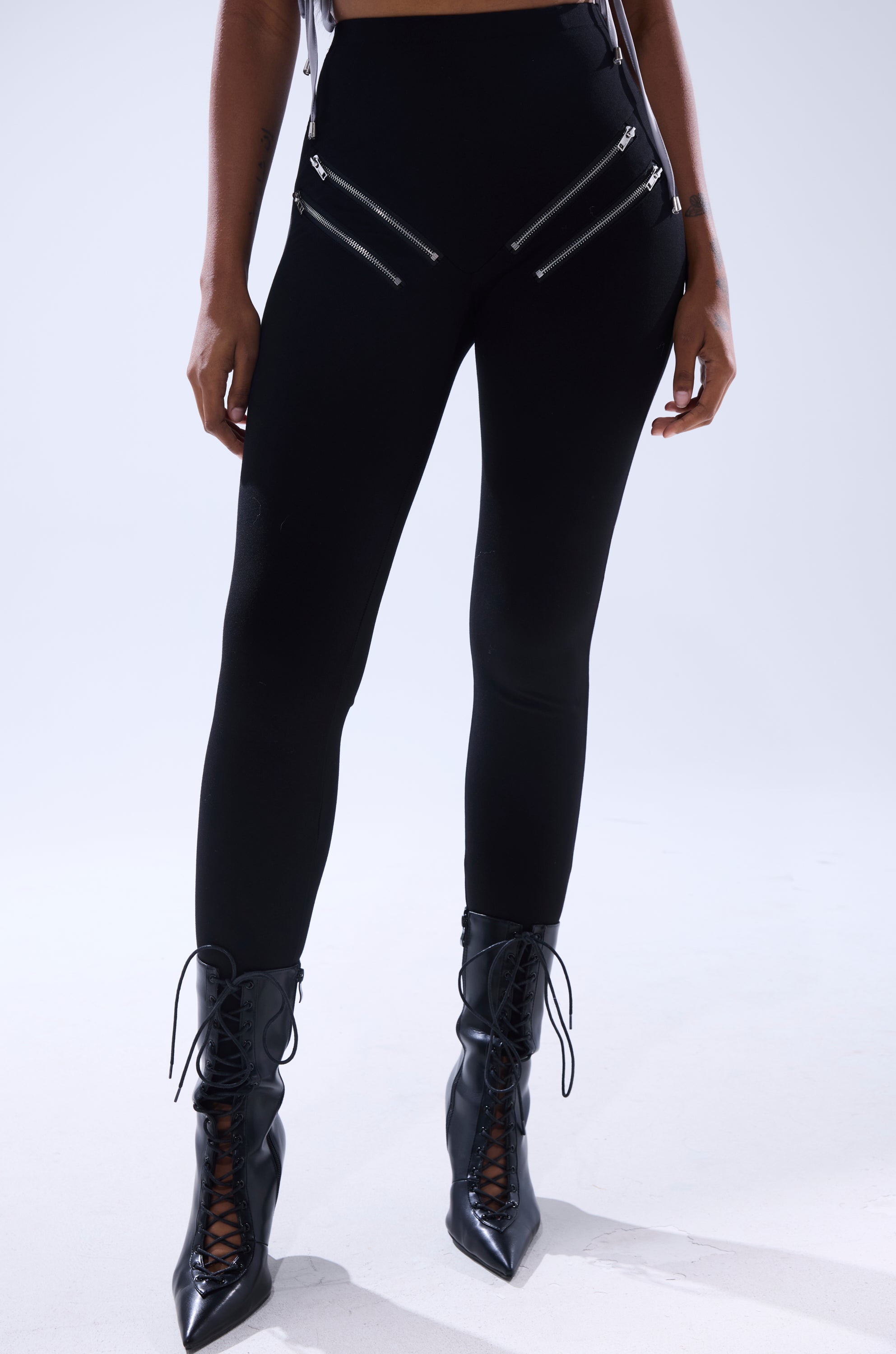 A person wearing ZIP ME UP SEAMLESS LEGGING, featuring high-rise black design with diagonal zippers, paired with black lace-up pointed-toe boots, stands against a plain light background.