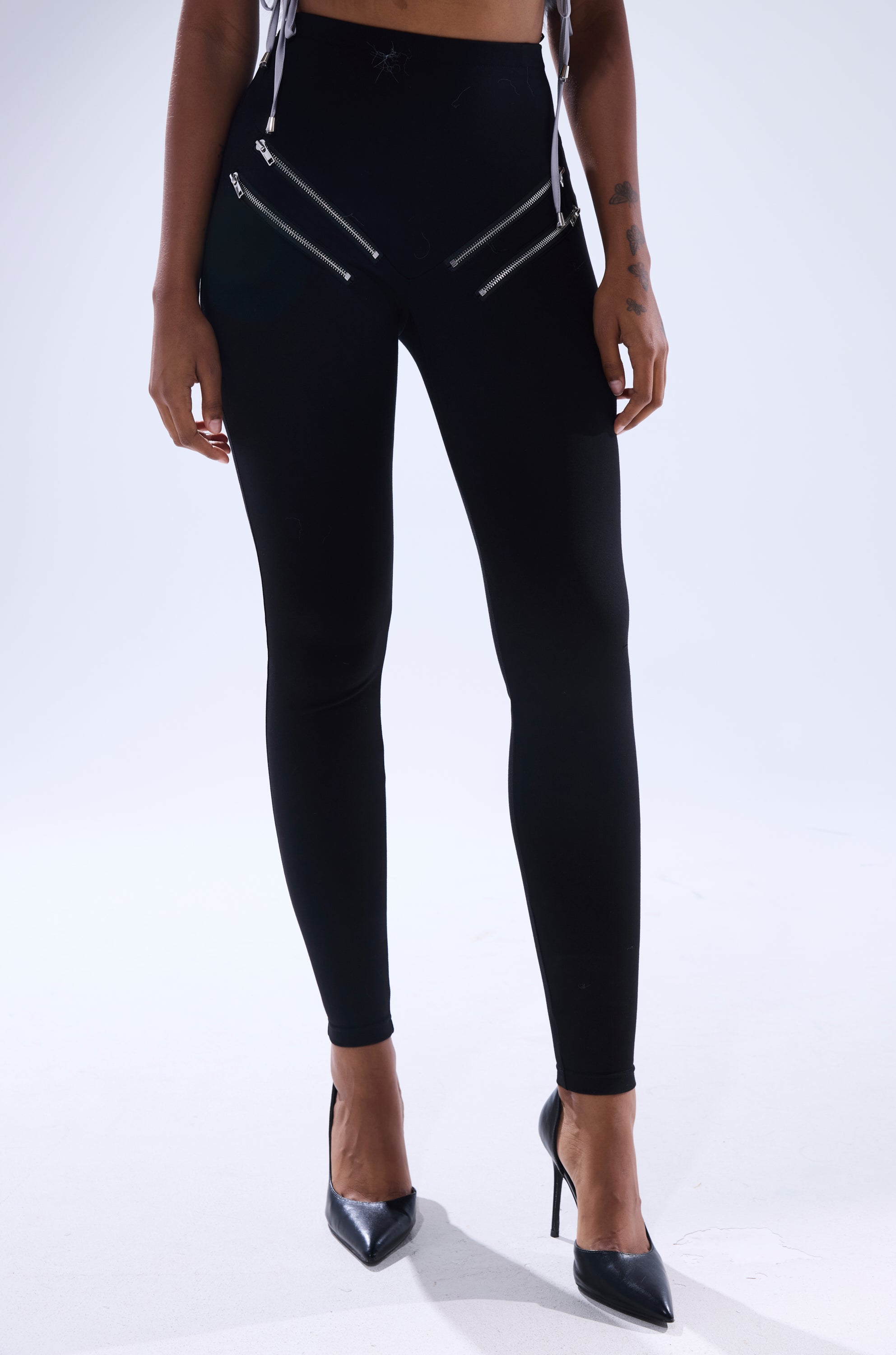 A person wearing the ZIP ME UP SEAMLESS LEGGING, featuring black high-rise design with two diagonal hip zippers, paired with black pointed-toe heels, stands against a white background.