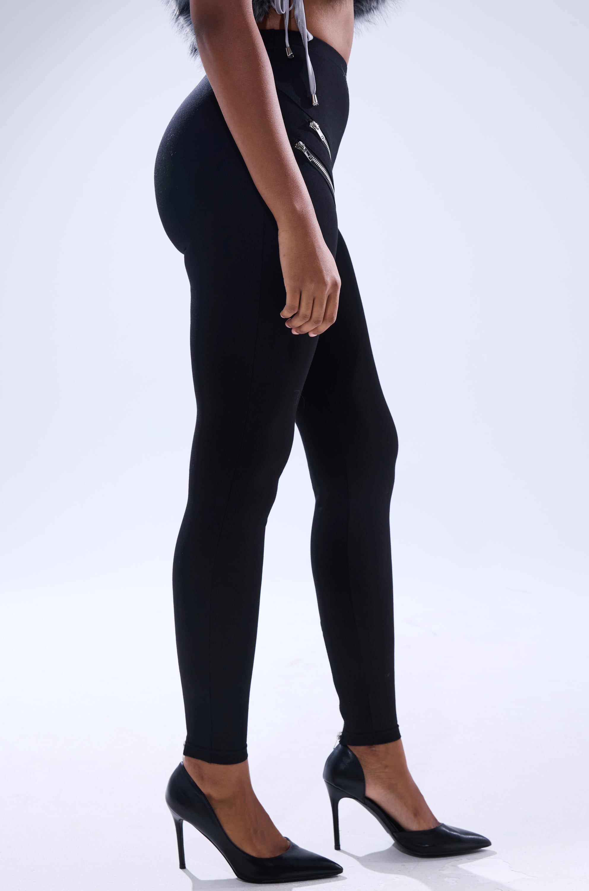 A model showcases the lower half of their body wearing ZIP ME UP SEAMLESS LEGGING in black with black high-heeled shoes against a plain white background.