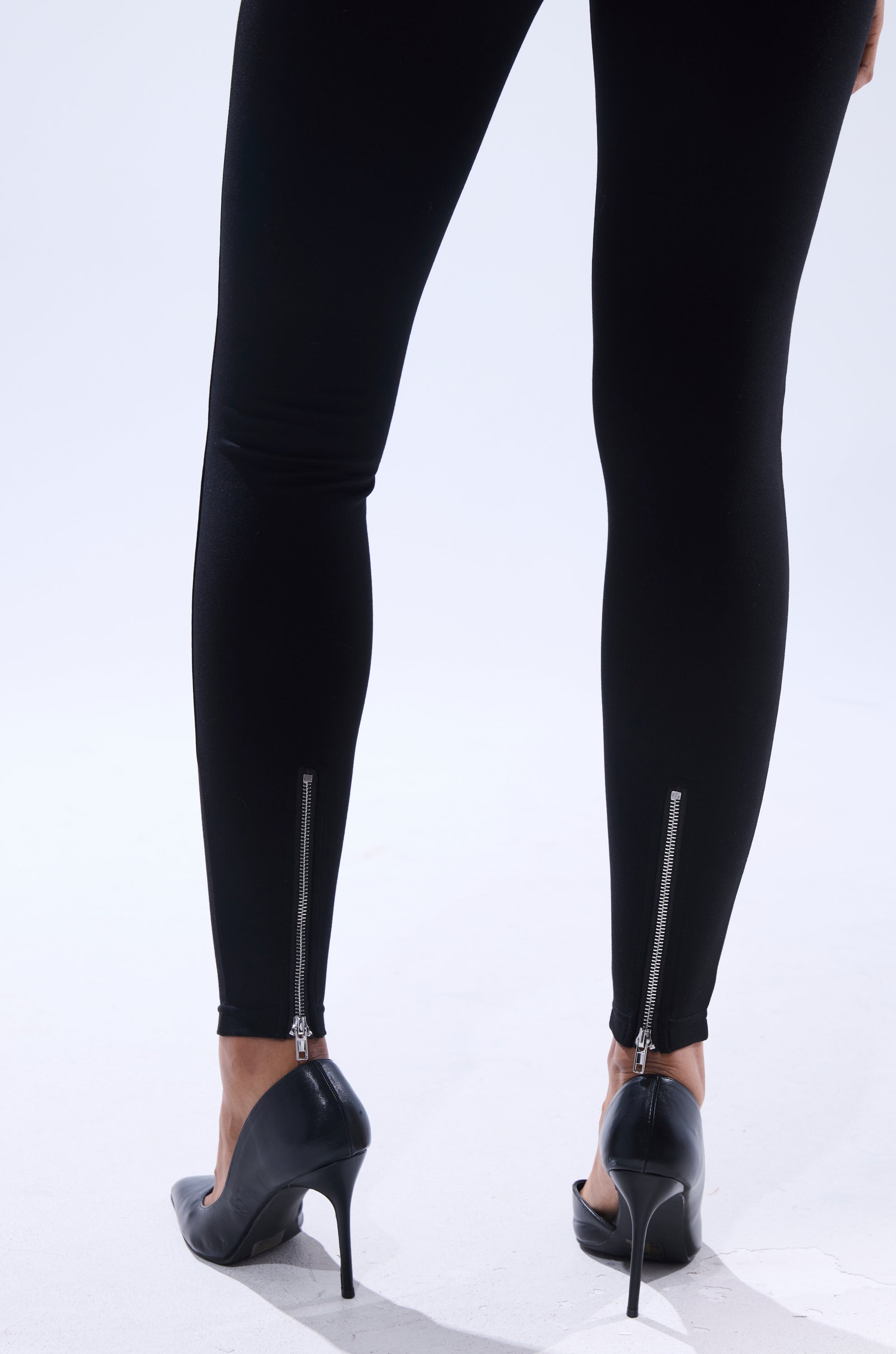 A person models the ZIP ME UP SEAMLESS LEGGING, featuring ankle zippers, paired with black high heels. Only the lower legs and feet are visible from behind, standing on a white surface.