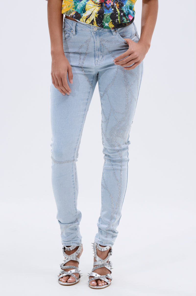 A person models the SPIDER GIRL SKINNY DENIM—light blue skinny jeans with rhinestone details—paired with a colorful top and metallic silver strappy high-heeled sandals against a plain white background. 0