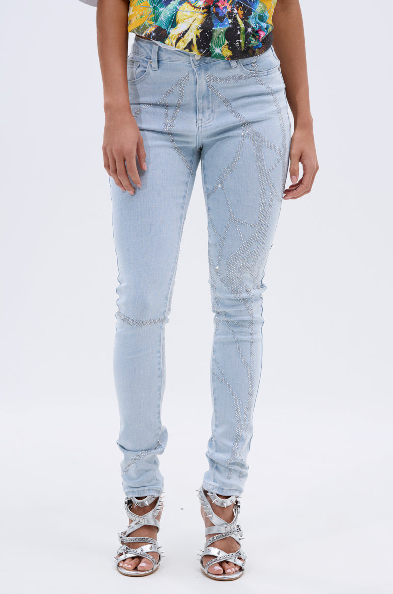 A person wearing SPIDER GIRL SKINNY DENIM in light blue with rhinestone details, a colorful patterned top, and silver strappy high-heeled sandals stands against a plain white background. 1