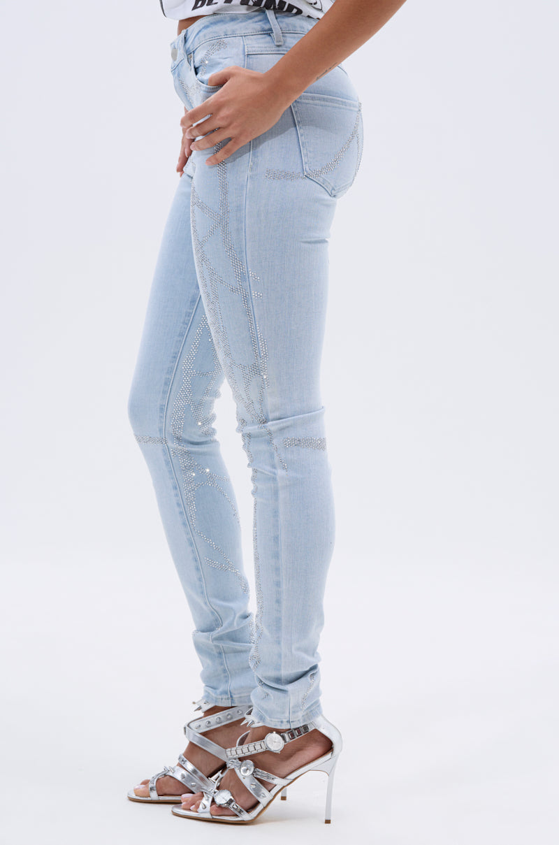 A woman poses sideways in SPIDER GIRL SKINNY DENIM, light blue skinny jeans with rhinestone details, paired with strappy silver high-heeled sandals against a plain white background. 3