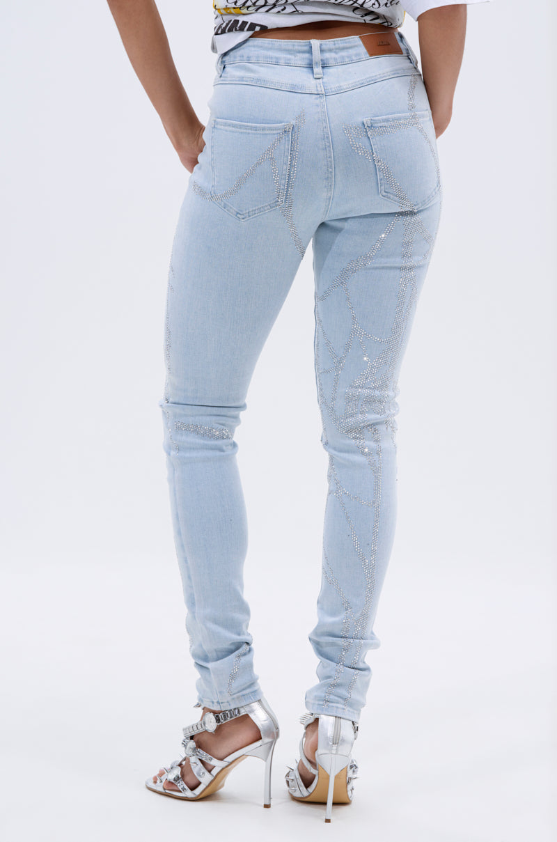 A person stands facing away against a white background, wearing SPIDER GIRL SKINNY DENIM—light-wash, high-rise skinny jeans with rhinestone-embellished back pockets—and silver high-heeled sandals. 4