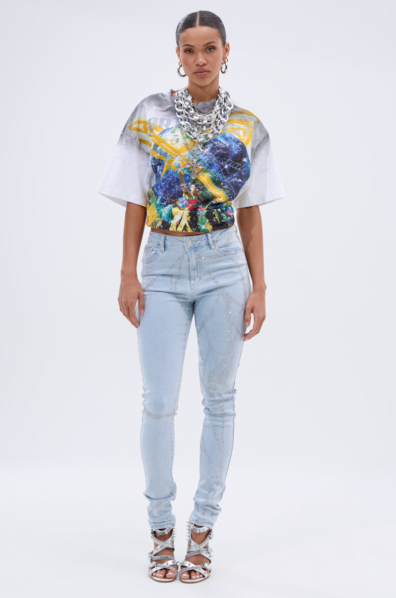 A woman stands against a plain background wearing a colorful graphic T-shirt, SPIDER GIRL SKINNY DENIM with rhinestone details, silver heels, chunky necklaces, and hoop earrings. 6