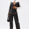 A person poses confidently against a plain white background wearing a stylish black and white cropped jacket, sheer patterned top, sunglasses, small black handbag, and the ROCK MY WORLD FAUX LEATHER LACE UP PANT.