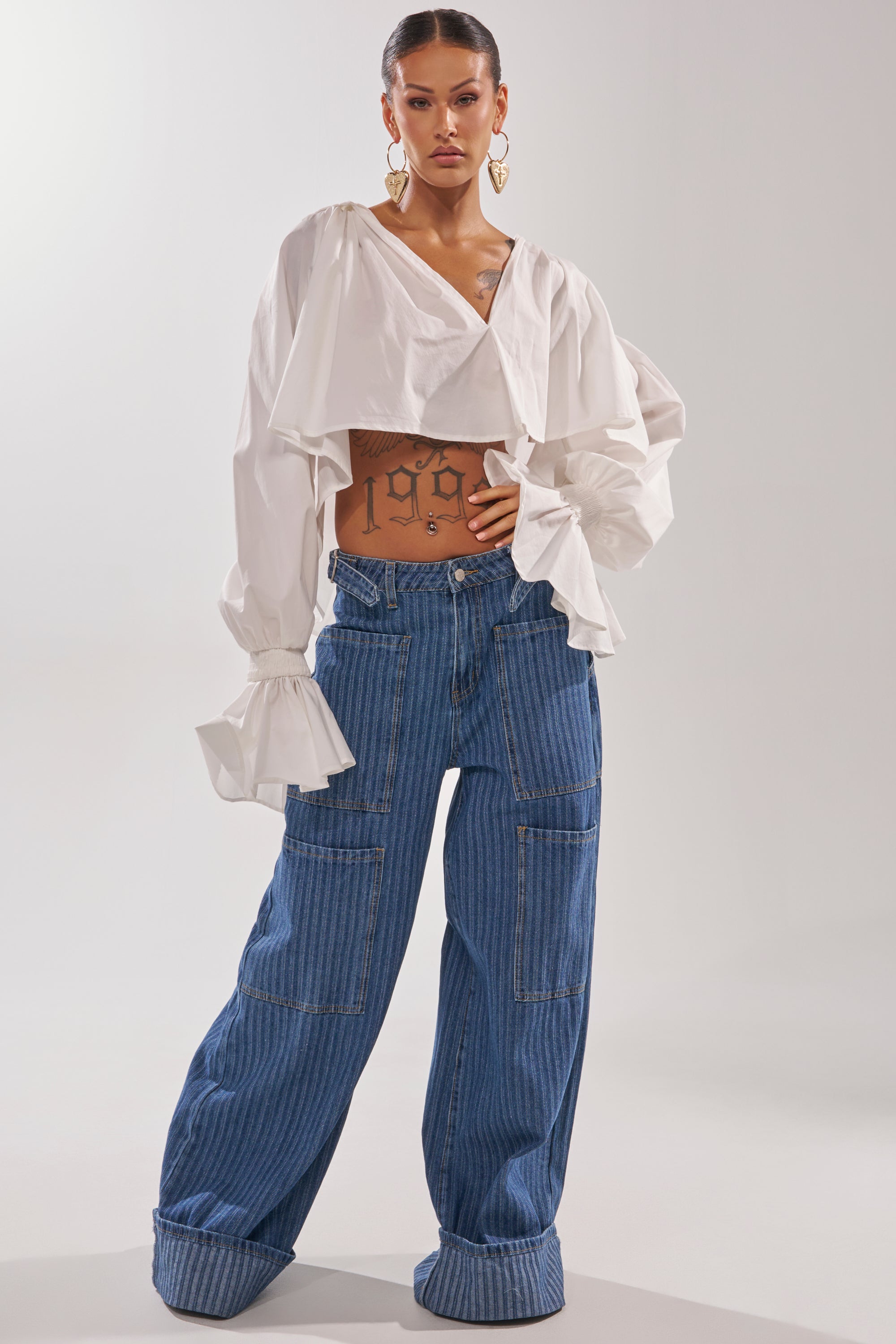 A woman with visible tattoos and large hoop earrings poses in a white, long-sleeve, cropped blouse with ruffled cuffs and high-rise BE MINE STRIPE DENIM wide-leg pants. 2