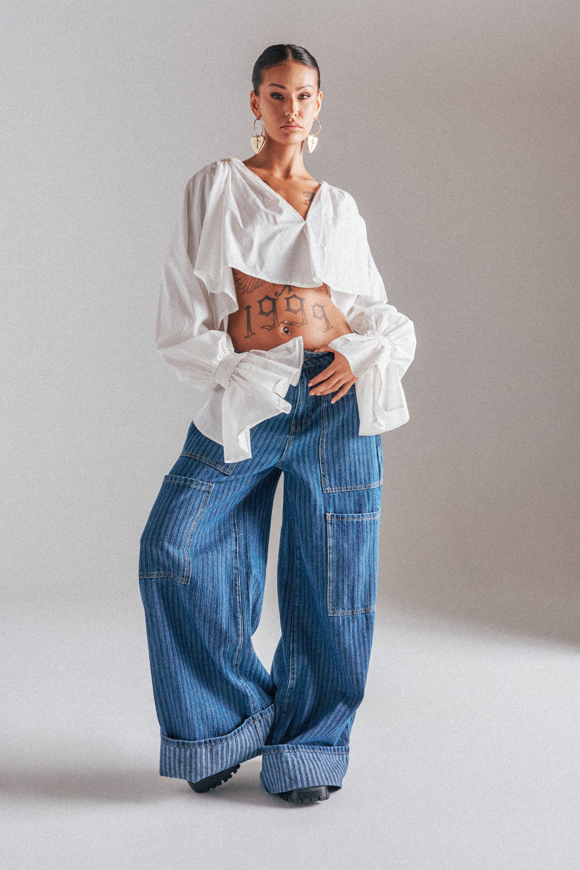A woman wears the BE MINE STRIPE DENIM wide-leg, high-rise jeans paired with a loose white blouse, showcasing a "1999" tattoo on her stomach. She accessorizes with hoop earrings and stands against a plain light background, hair tied back. 1