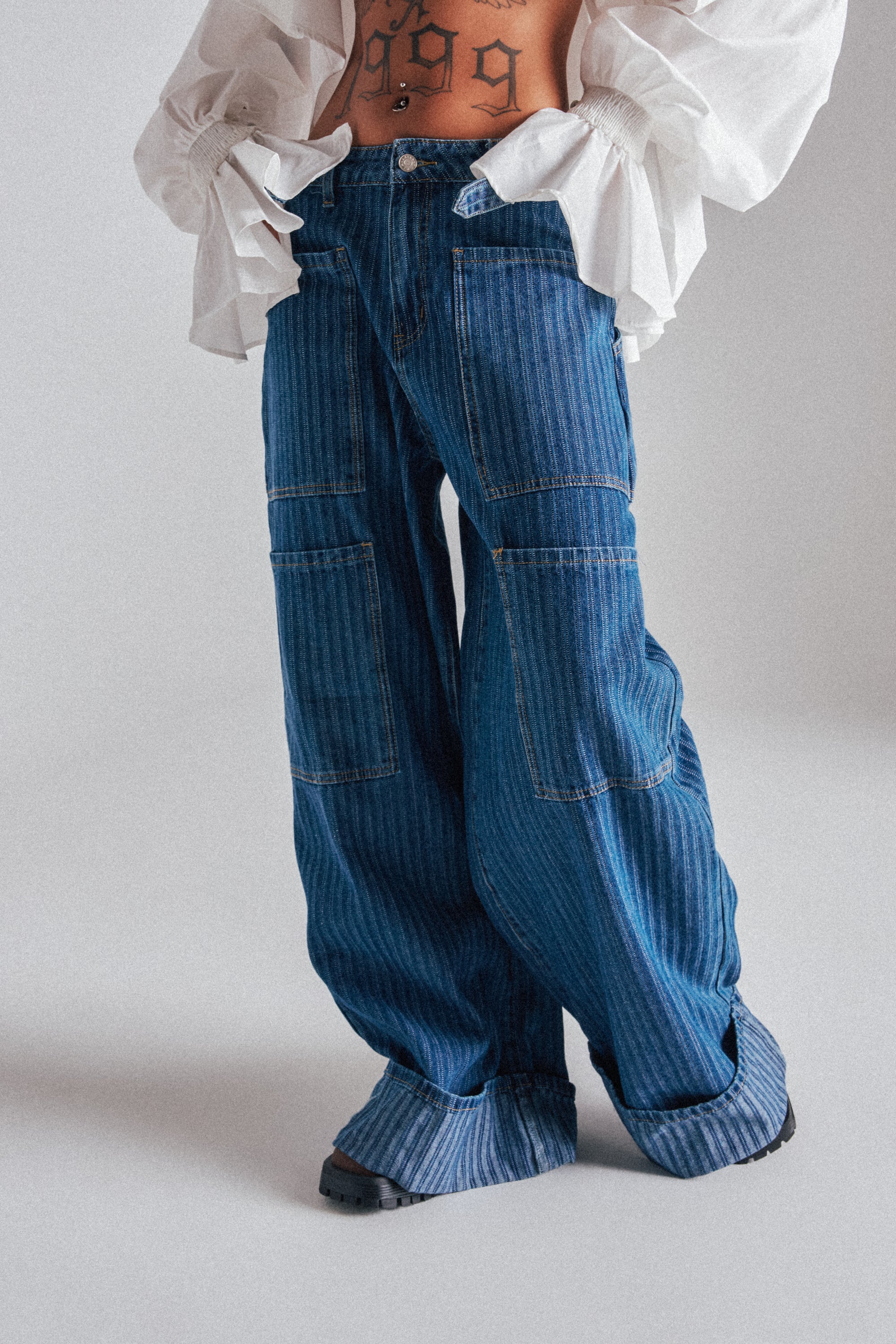 A person wears BE MINE STRIPE DENIM, featuring loose, wide-leg dark wash blue jeans with visible patches, paired with black shoes and a white voluminous-sleeved blouse. A tattoo shows on their exposed midriff against a plain, light background. 0