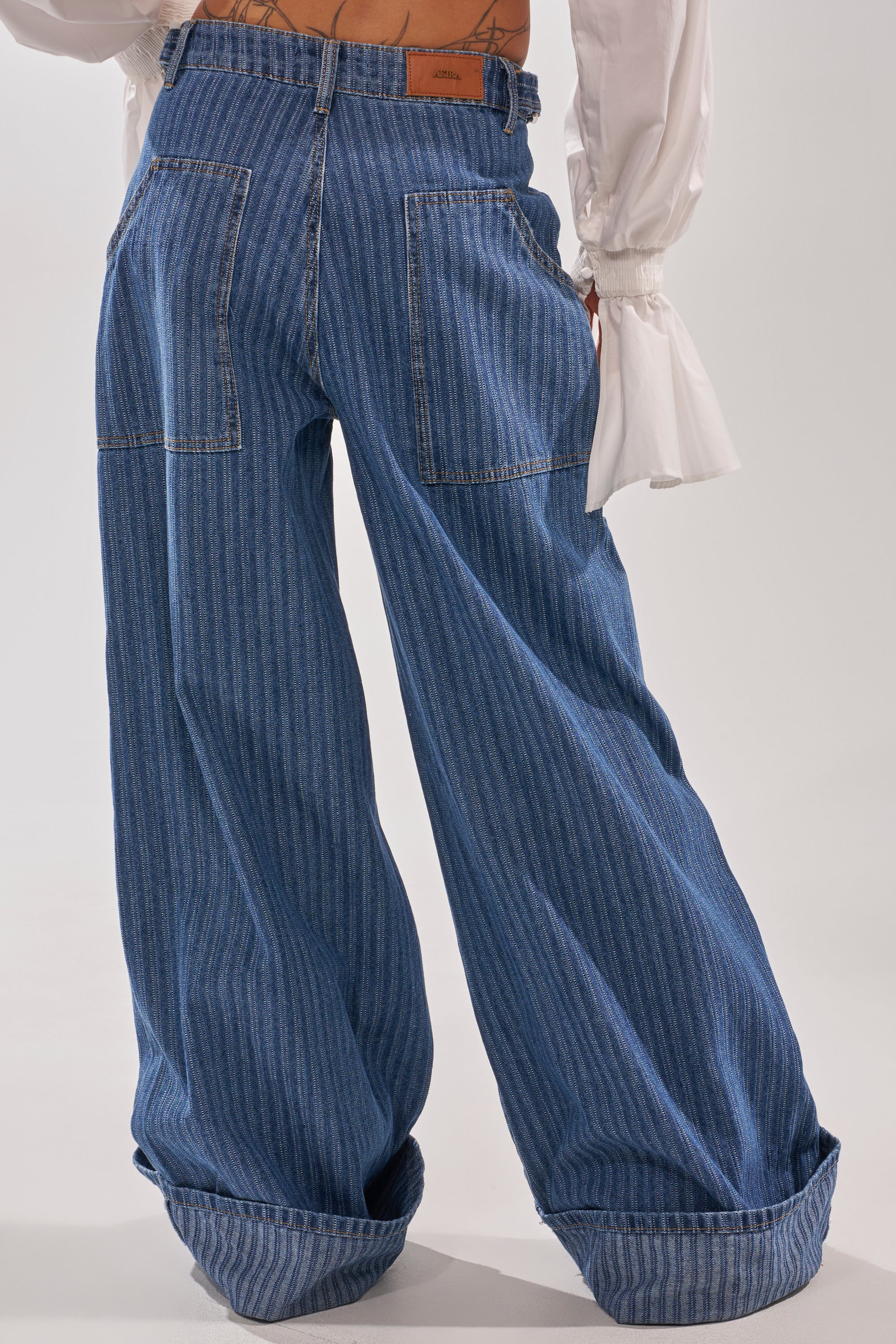 Person wears the BE MINE STRIPE DENIM: high-rise, wide-leg pants with blue pinstripes, large back pockets, and cuffed hems. Paired with a white blouse featuring ruffled sleeves. Image highlights lower back and legs. 5