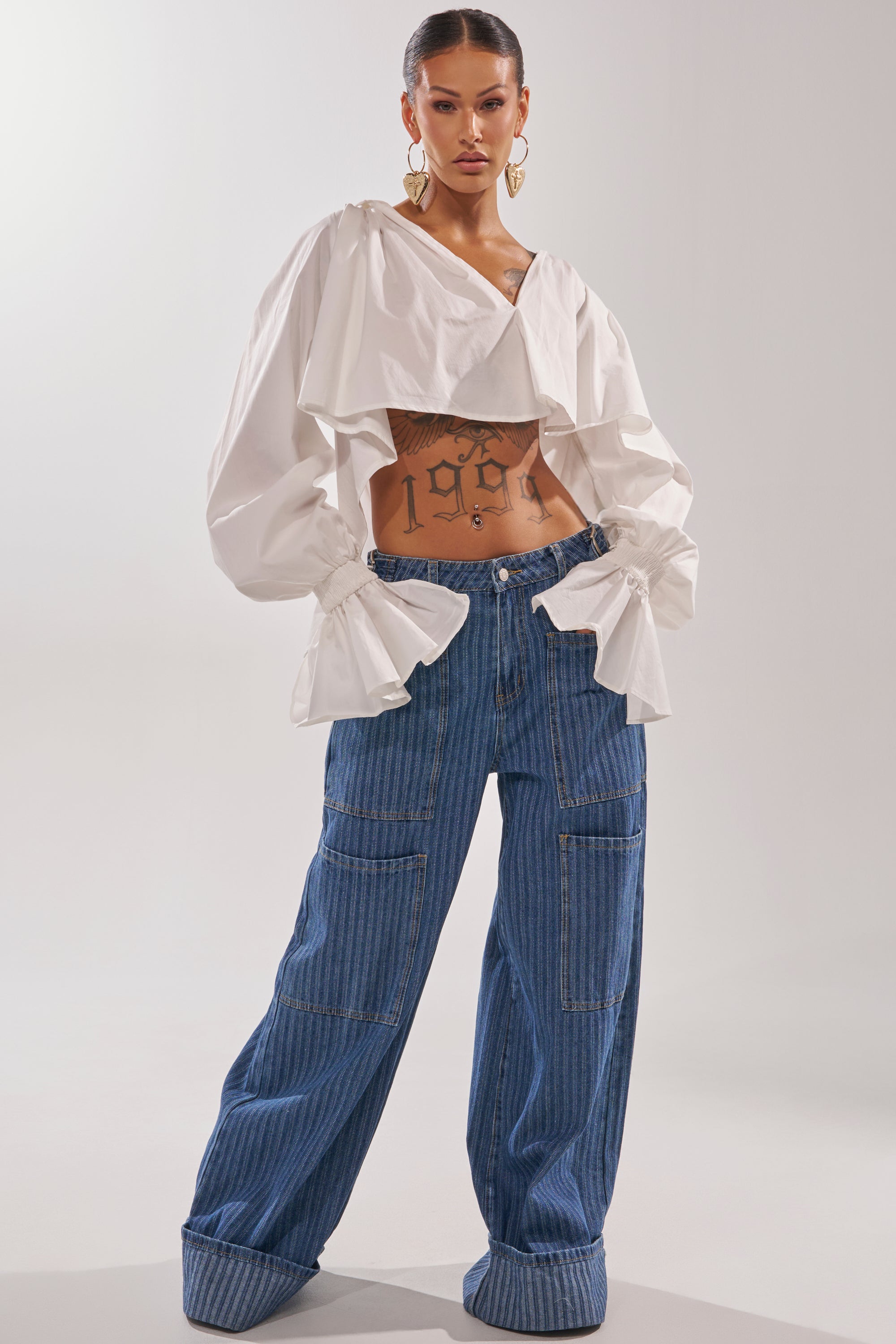 A woman poses confidently in the BE MINE STRIPE DENIM, wide-leg high-rise jeans with large cuffs, styled with a cropped white blouse, gold hoop earrings, slicked-back hair, and a "1998" tattoo visible on her torso. 6