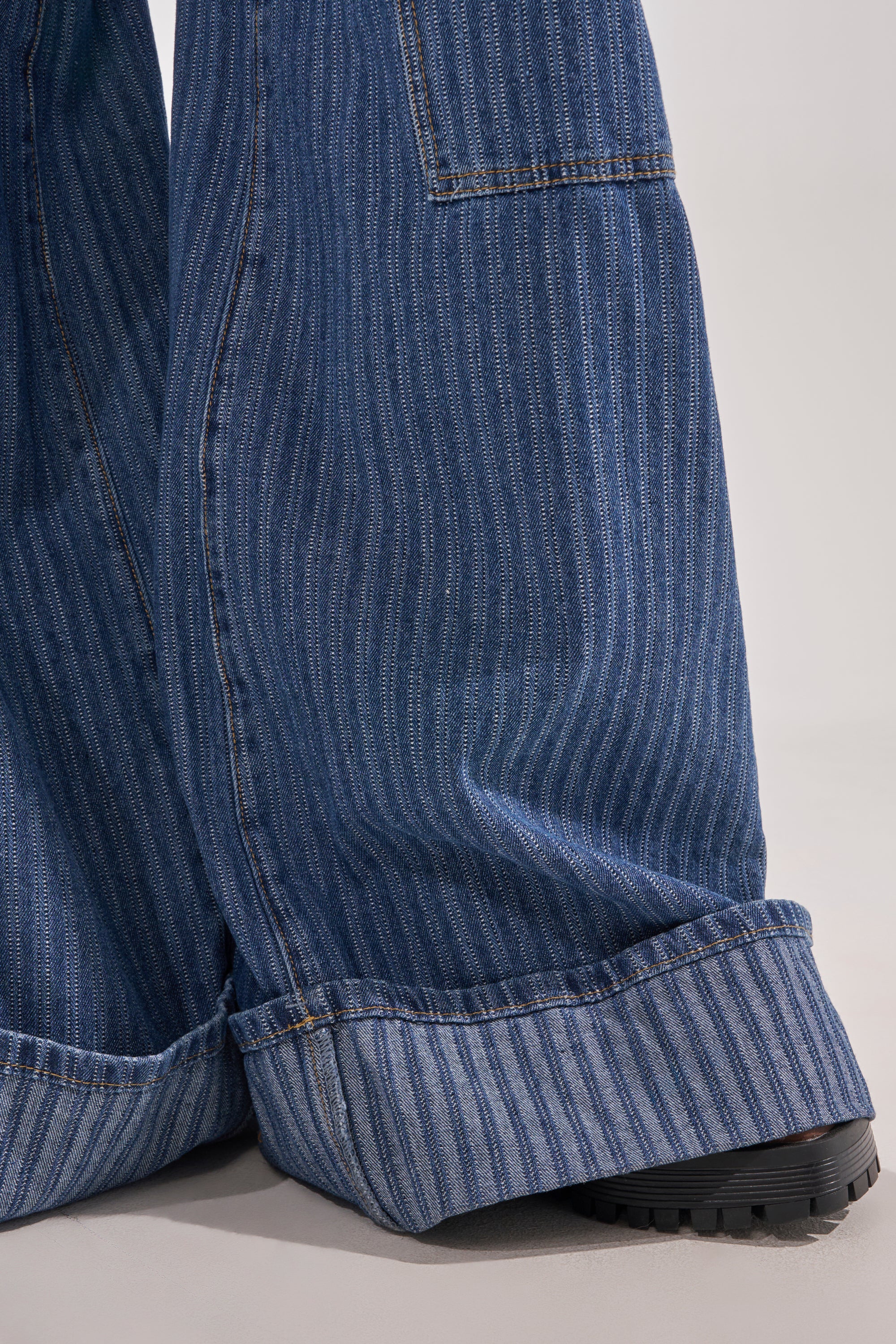 Close-up of lower legs wearing BE MINE STRIPE DENIM high-rise, wide-leg pants with blue pinstripes and large cuffs, partially covering black shoes on a light-colored floor. 8