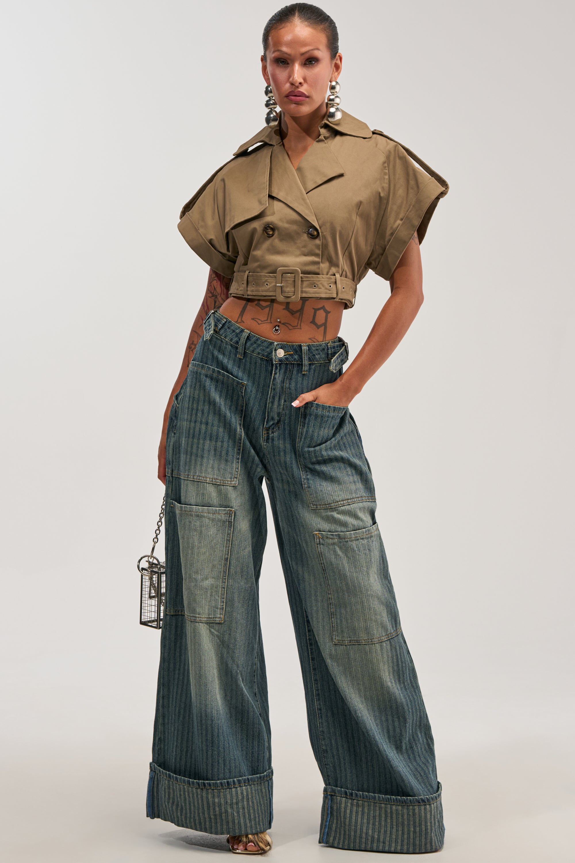 A woman poses in a cropped, khaki belted jacket with wide sleeves, BE MINE STRIPE DENIM high-waisted streetwear jeans featuring large cuffs and pockets, sandals, bold earrings, and a small metallic purse.
