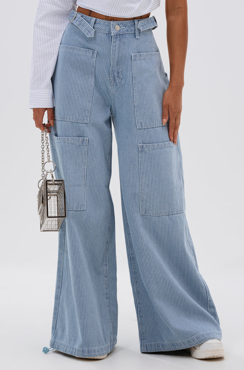 Only the lower body is visible: someone in SUMMERTIME BLUES STRIPE WIDE LEG DENIM pants with patch pockets and white shoes holds a small, silver, box-shaped metal handbag. 2
