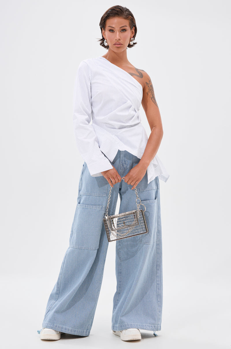 A person with short hair wears a white, asymmetrical, one-shoulder blouse and SUMMERTIME BLUES STRIPE WIDE LEG DENIM. They hold a small silver handbag and wear white shoes, standing against a plain white background. 6