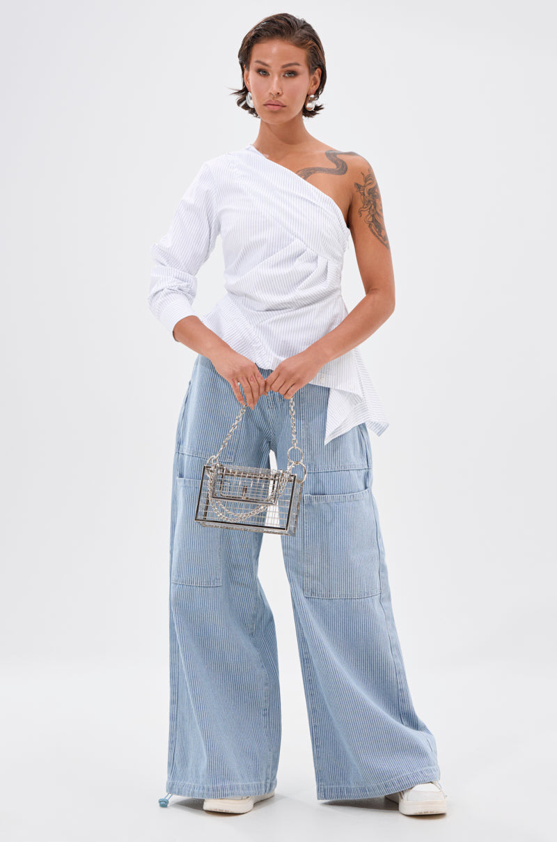 A person stands confidently against a plain background, wearing a one-shoulder white blouse, SUMMERTIME BLUES STRIPE WIDE LEG DENIM in light blue, white shoes, and holding a small transparent chain-strap handbag. 7
