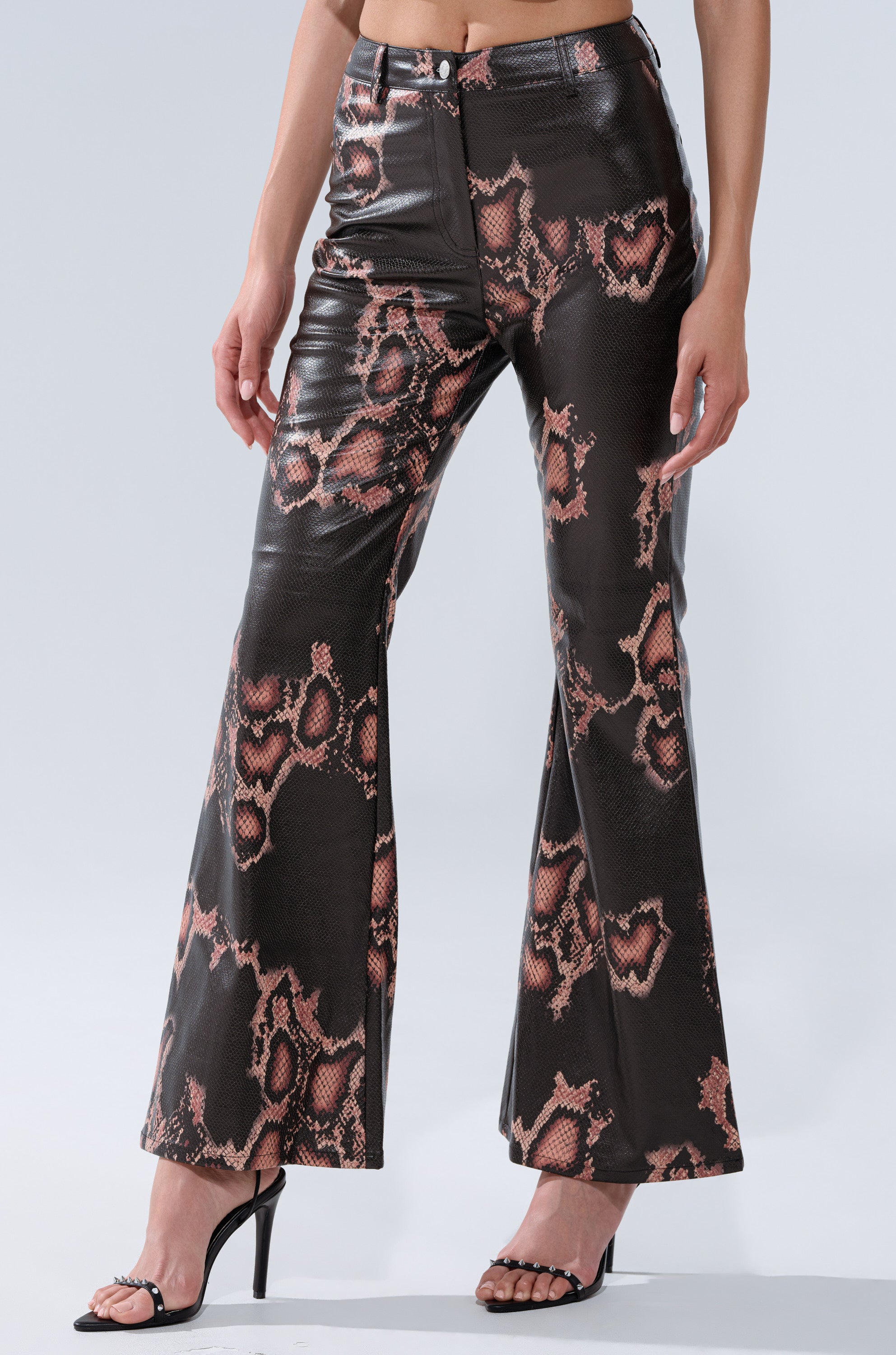 A person wears COLD BLOODED FAUX LEATHER PANTS, which are high-waisted with a brown and pink snakeskin print and flared legs, along with black heeled sandals. Their midriff and lower legs are visible against a plain light background.