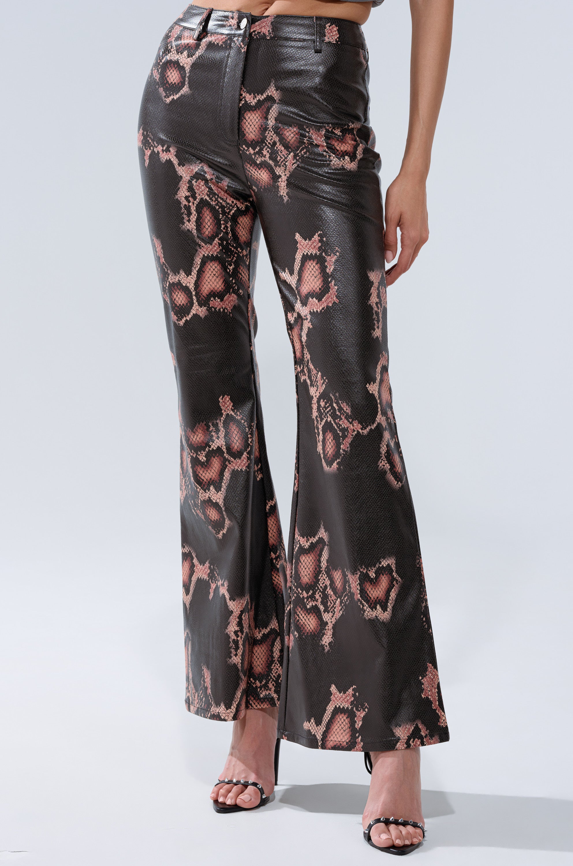 A person wearing COLD BLOODED FAUX LEATHER PANTS with a brown and pink snakeskin print and black heeled sandals stands against a plain light background.