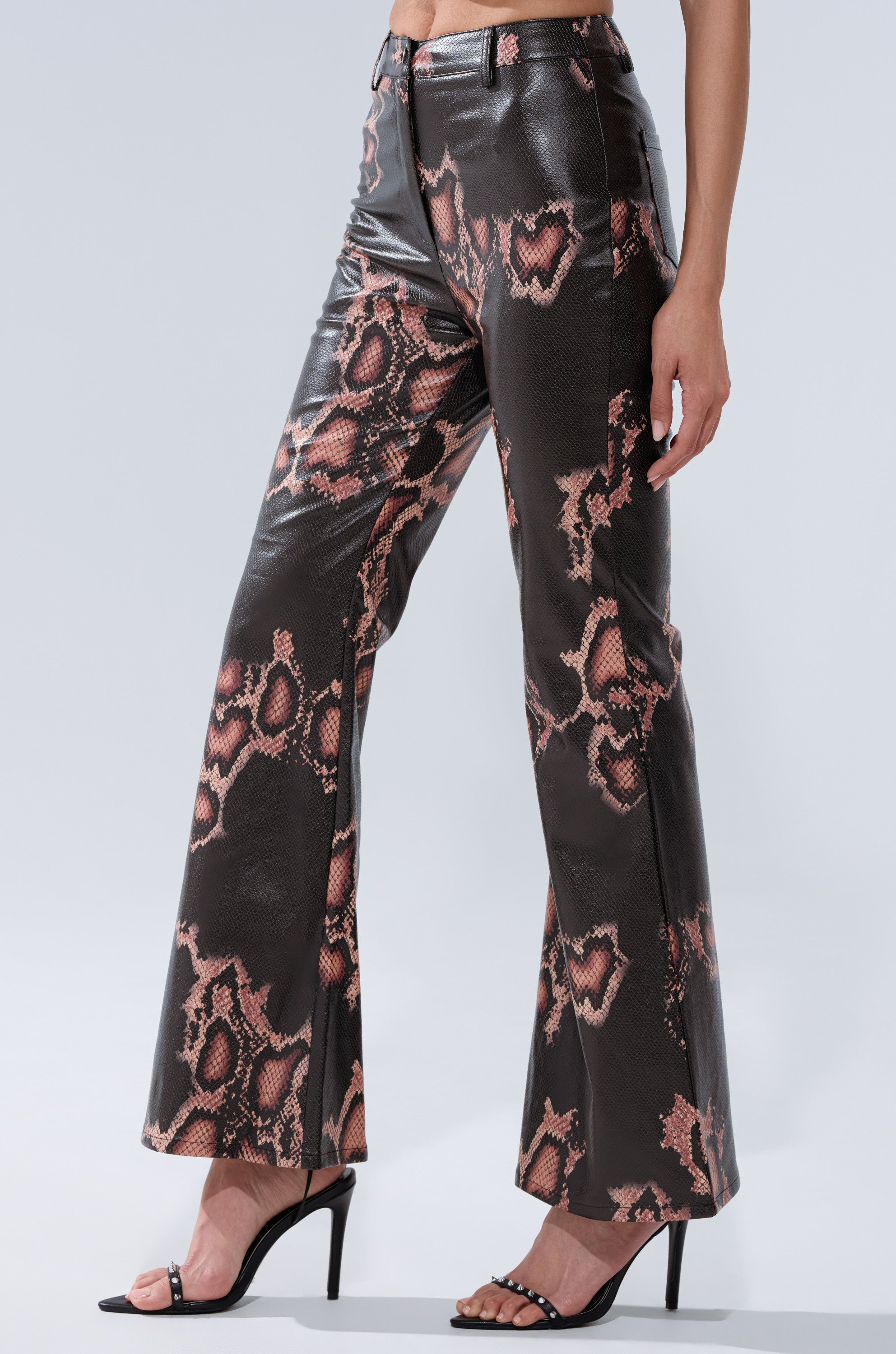 A person stands against a plain light background wearing COLD BLOODED FAUX LEATHER PANTS—high-rise, flared, brown and pink snakeskin print pants with wide legs—paired with black high-heeled sandals.